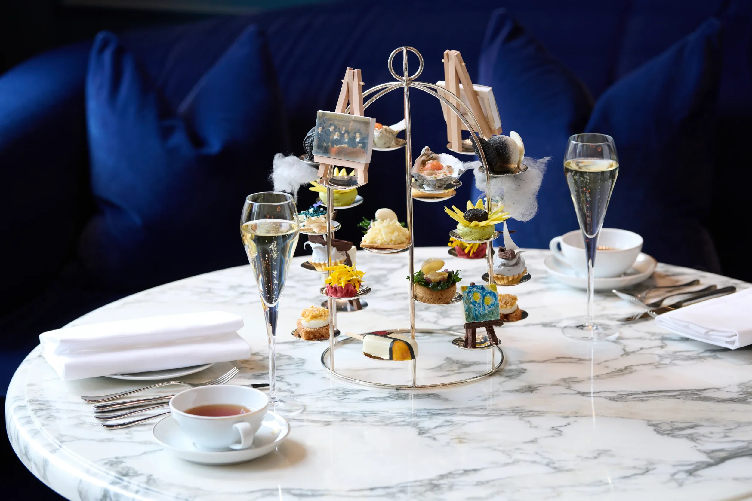 Elegant table setting with a tiered tray of assorted decorative desserts on a white marble table, flanked by champagne glasses, a cup of tea, and a white napkin, against a dark blue upholstered background.