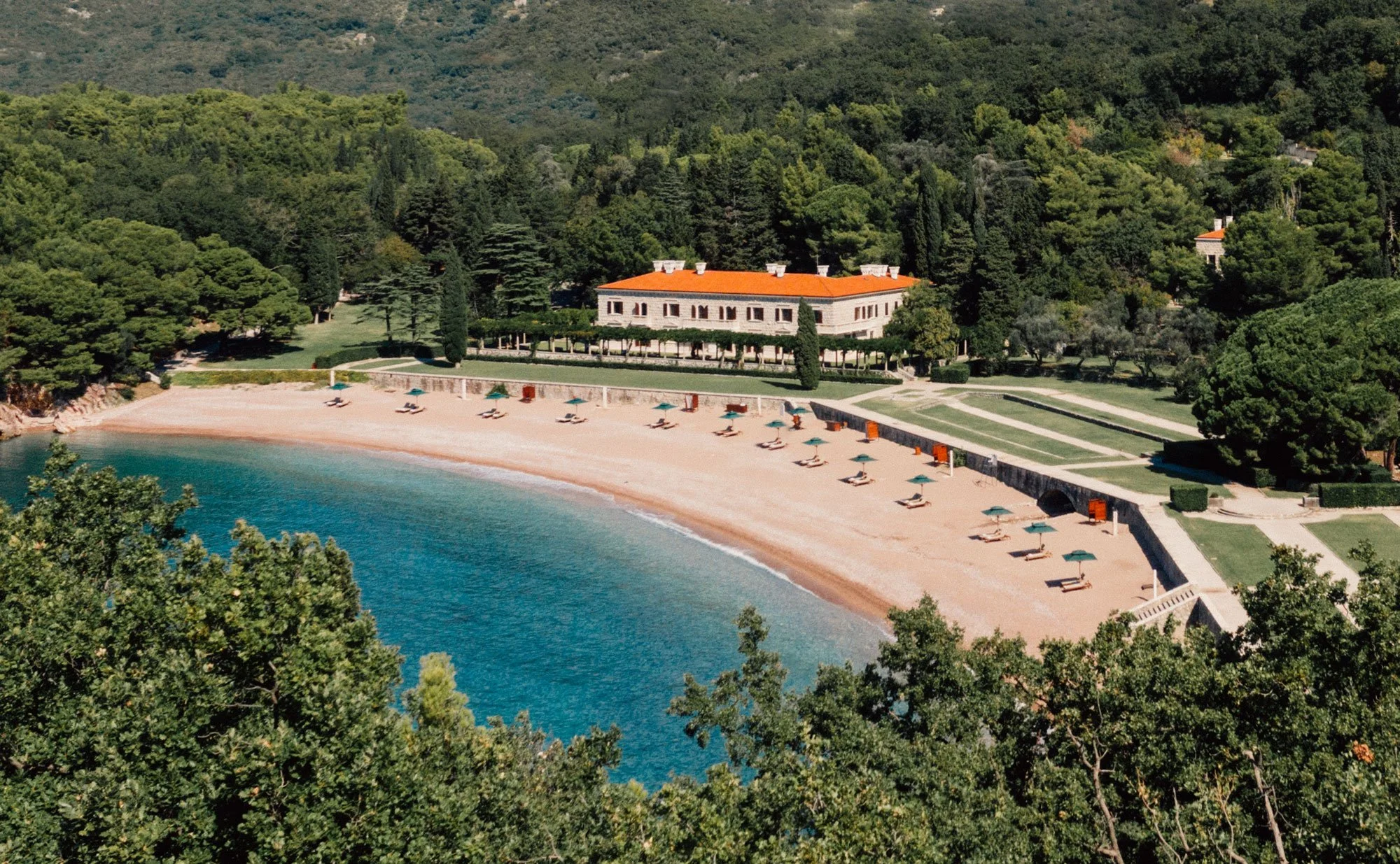 A scenic view of a private beach with lounge chairs and umbrellas, a large house on a hill surrounded by lush green trees, and a calm blue sea.