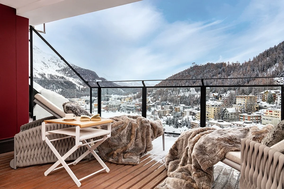 A cozy balcony with beige furniture and fur blankets overlooking a snow-covered mountain town.