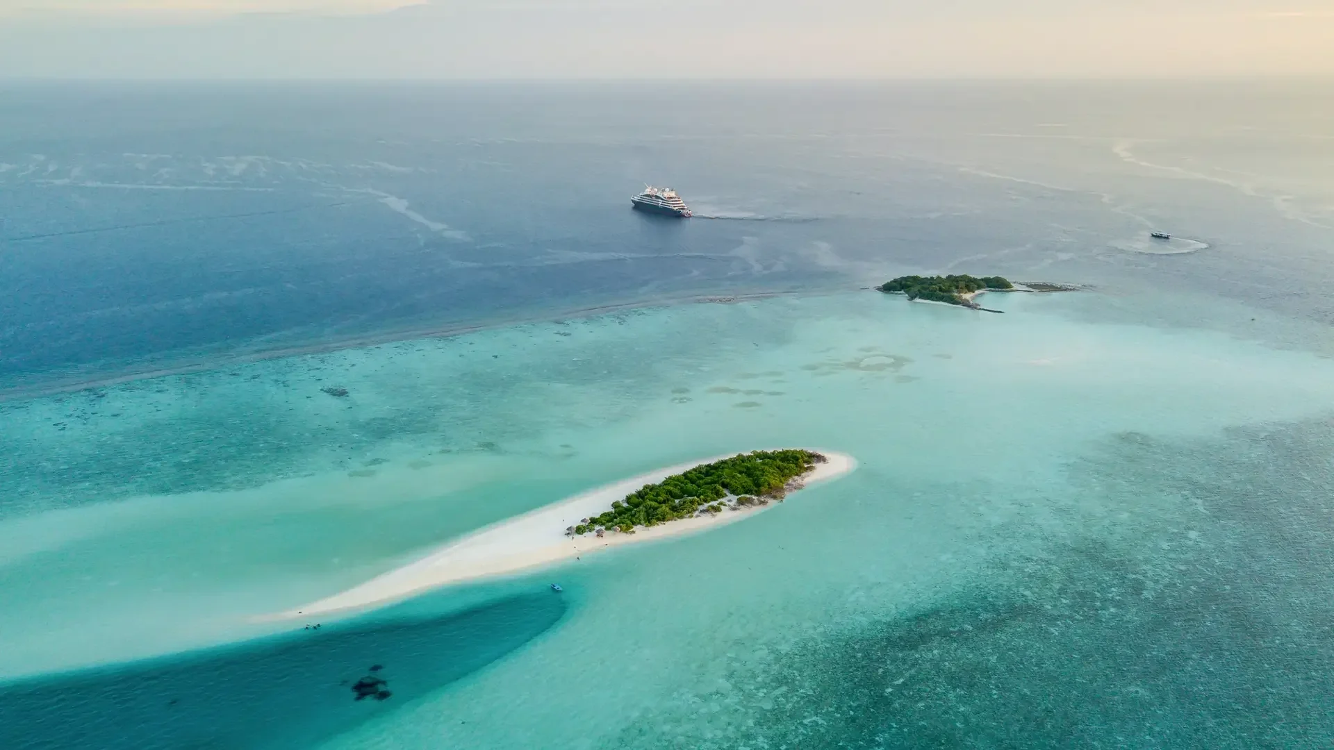 An aerial view of a small green island with white sandy beaches surrounded by turquoise water, with cruise ships in the distance.