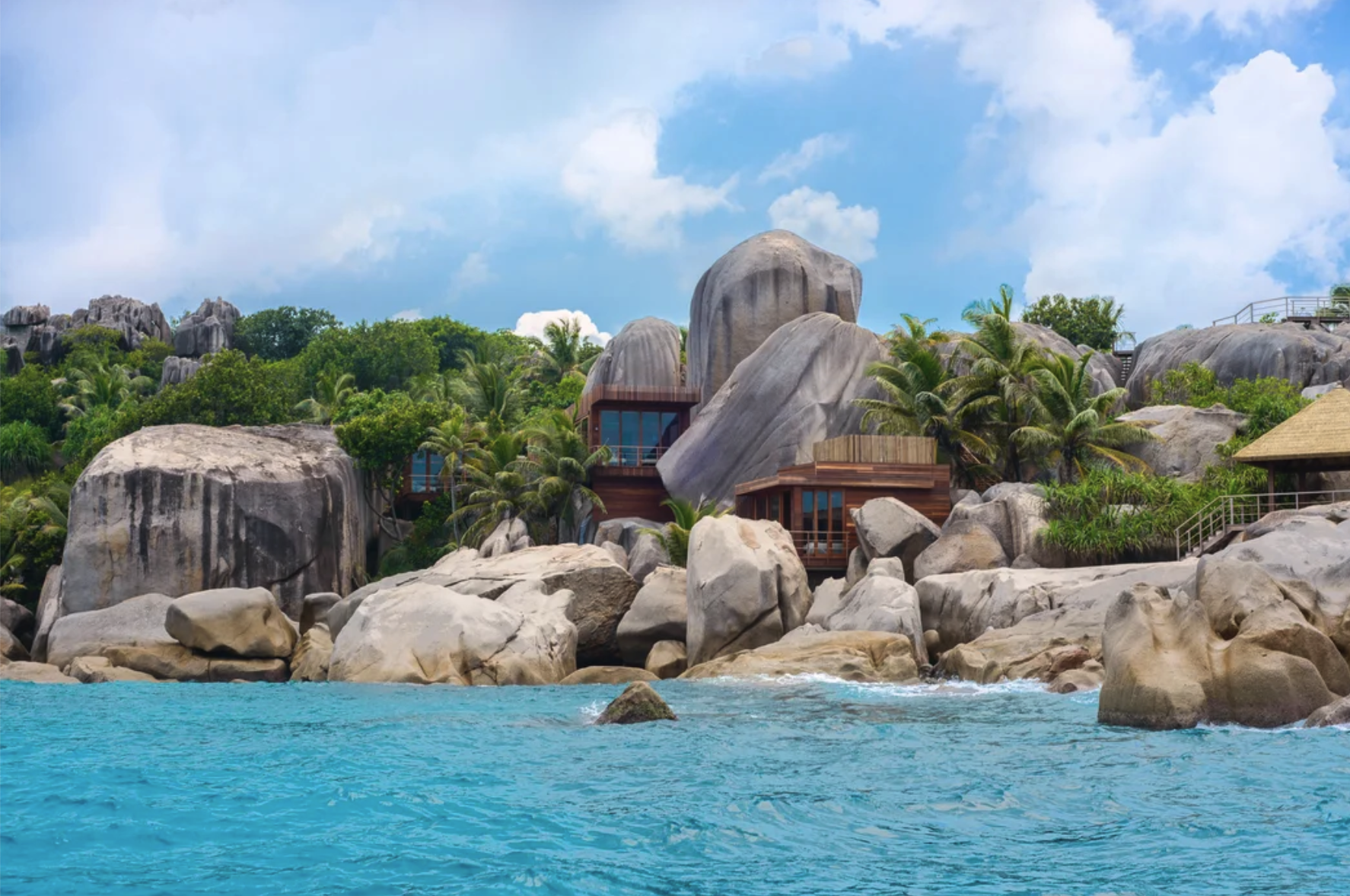 Tropical coastal scenery with large rocks, lush green palm trees, and wooden buildings on rocky terrain under a blue sky with clouds.