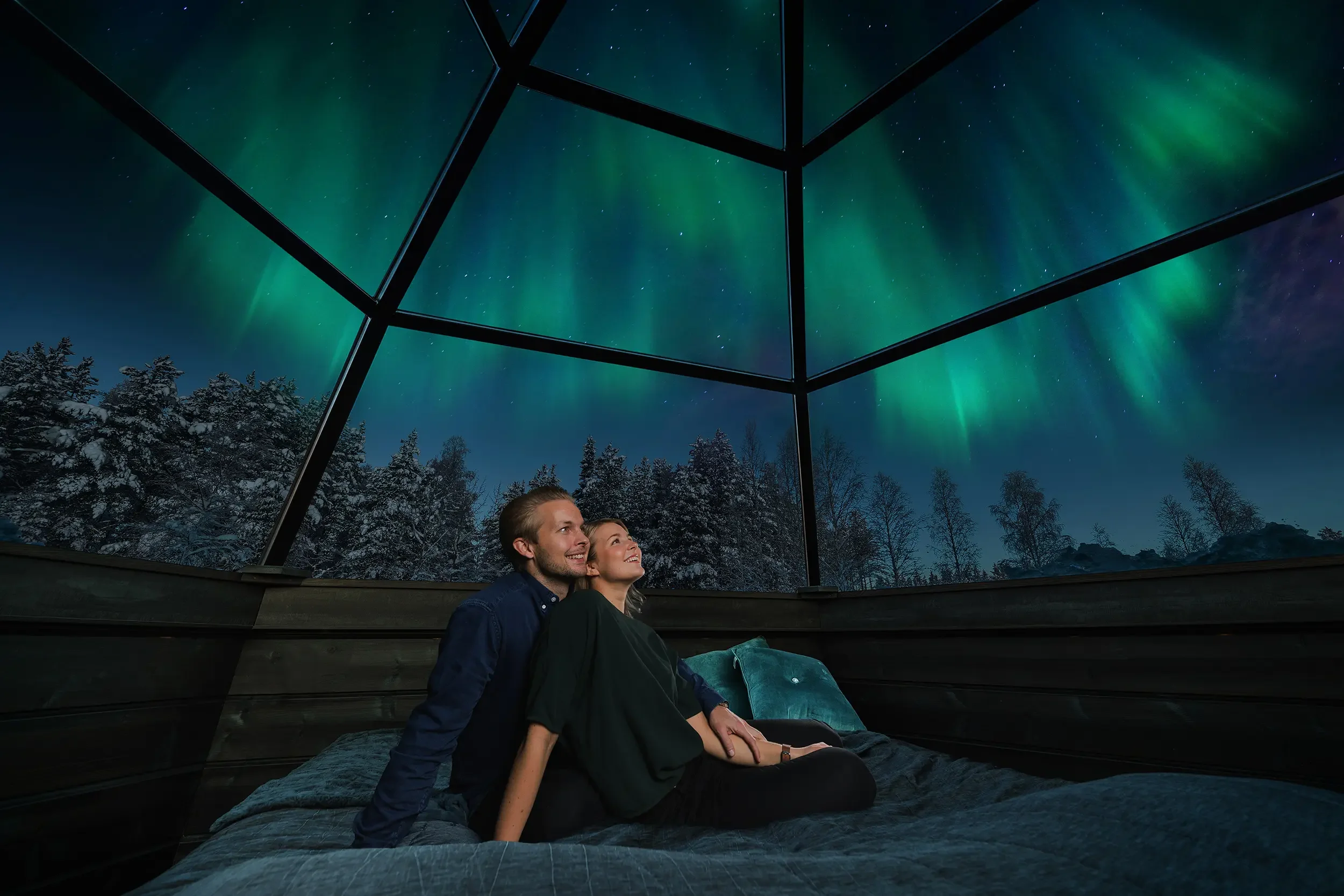 Couple sitting on bed inside a glass cabin, gazing up at the northern lights in the starry night sky