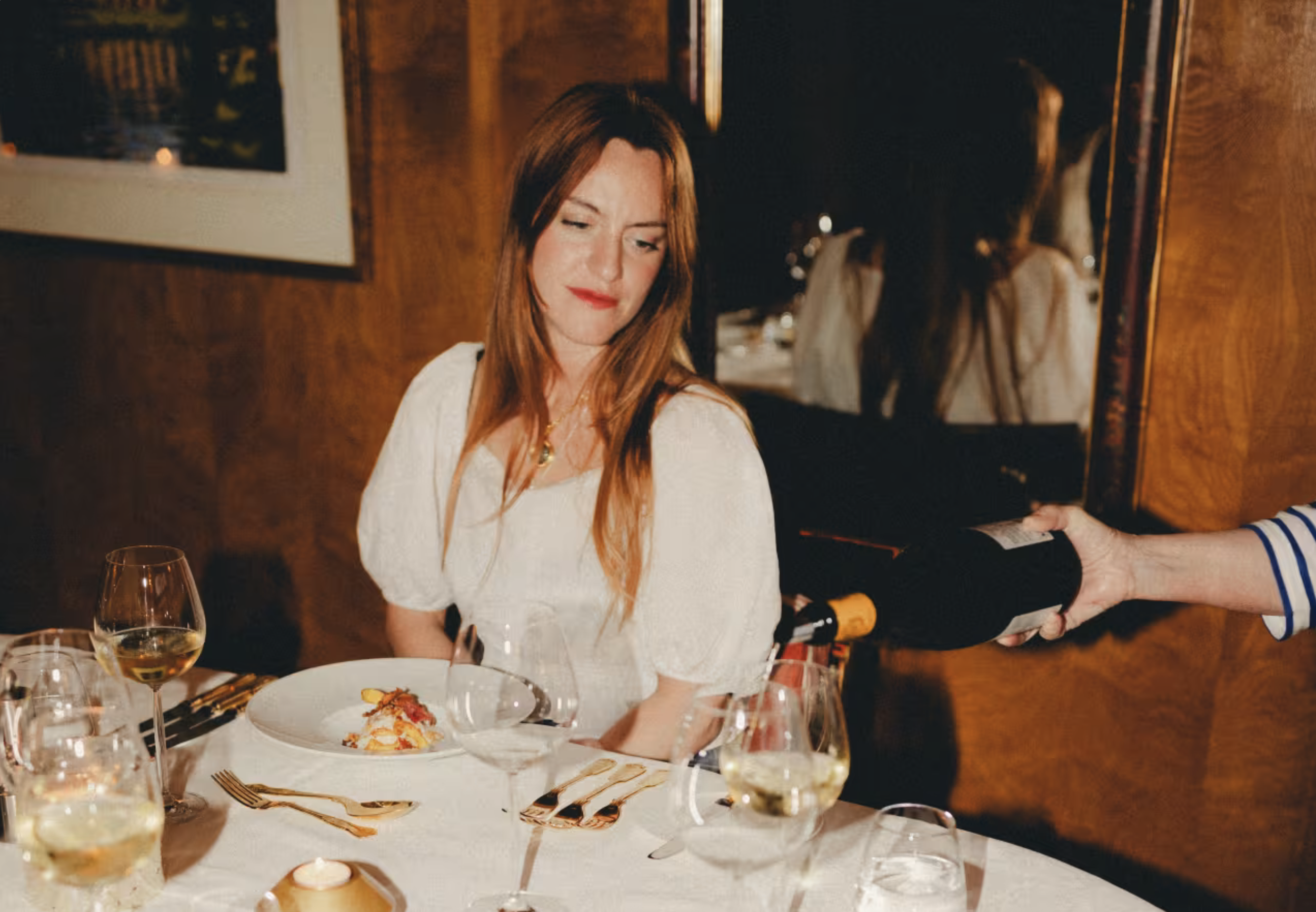 A woman with long red hair and a white dress sitting at a dining table, being served a glass of white wine in a restaurant with wooden wall decor and framed pictures.