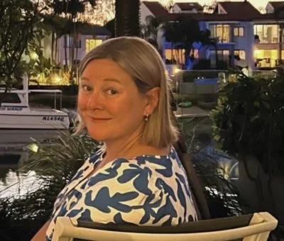 A woman with short blonde hair smiling at the camera, sitting outdoors near a body of water with boats and buildings in the background.