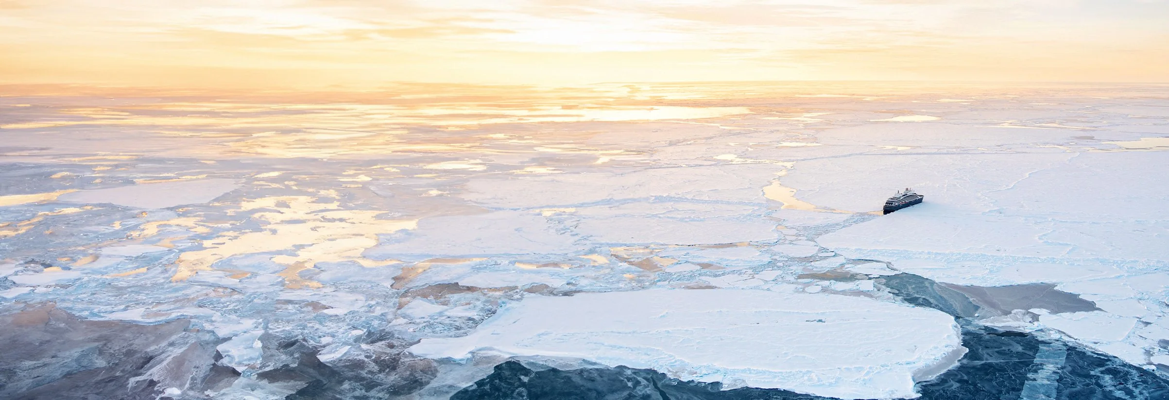 A ship navigating through icy, broken sea ice during sunset or sunrise.