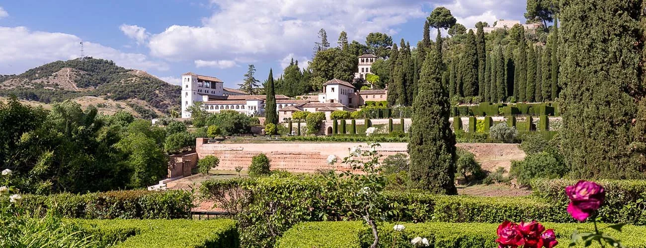 A lush, green hillside garden with multiple levels, tall cypress trees, and white buildings with terracotta roofs in the background. Pink roses are visible in the foreground, and the sky is partly cloudy.