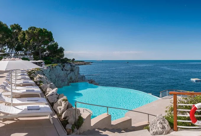 Infinity pool overlooking the ocean with lounge chairs and umbrellas on the left, rocky landscape, and a clear blue sky.