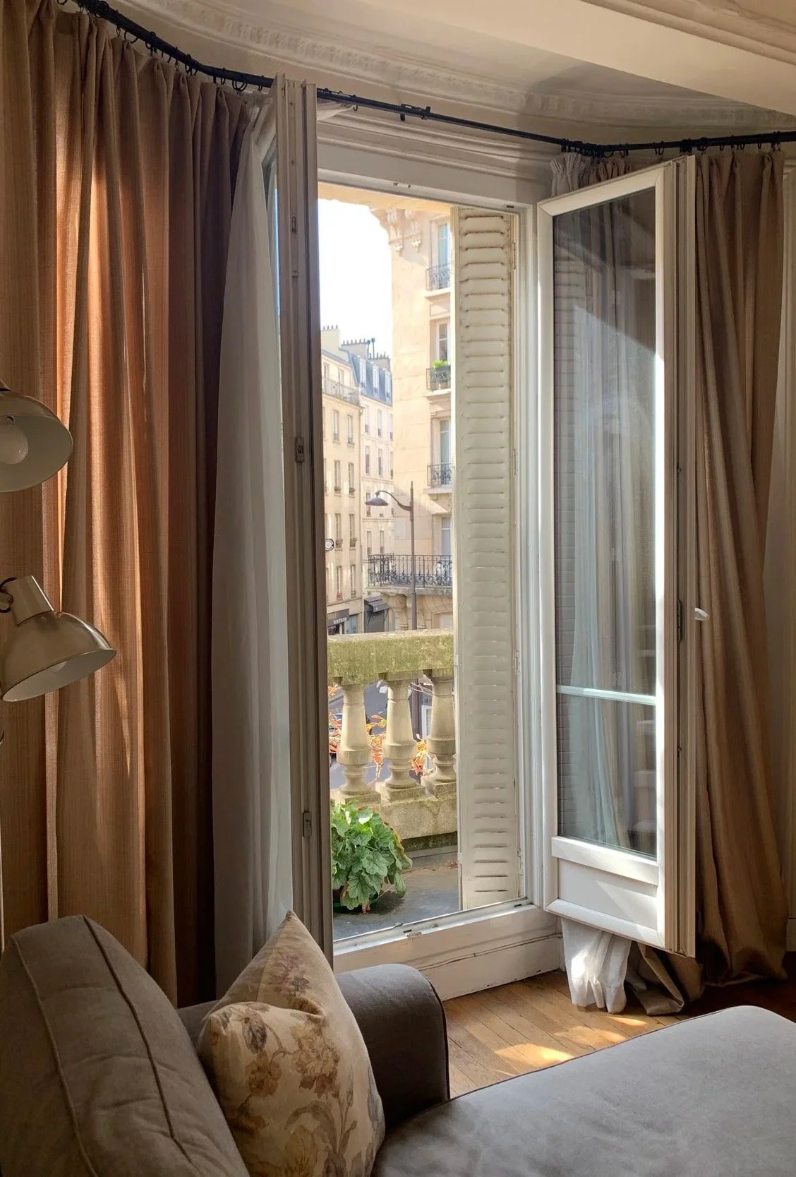 Open balcony door with French style shutters, beige curtains, and view of neighboring apartment buildings with balconies and street lamps.