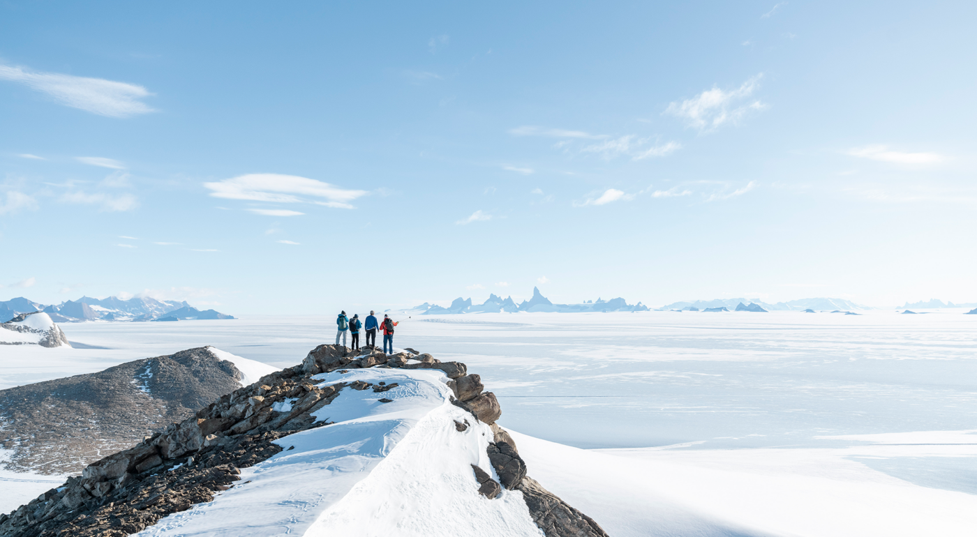 Four people in winter gear standing on a snowy mountain ridge overlooking a vast icy landscape with distant mountains under a clear blue sky.