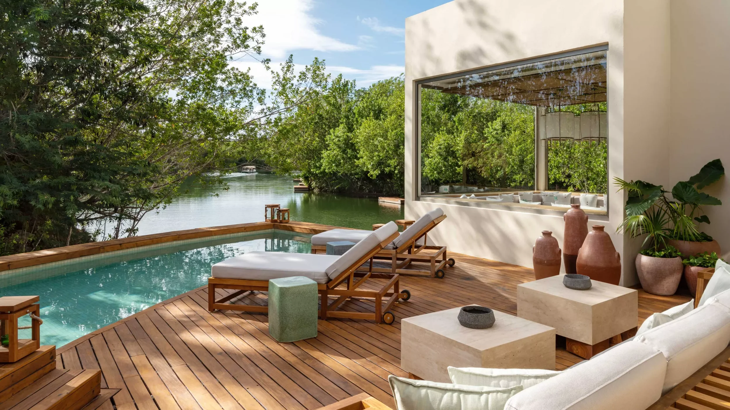 Luxury outdoor patio with a small pool overlooking a river, surrounded by trees, featuring lounge chairs, potted plants, and a large window.