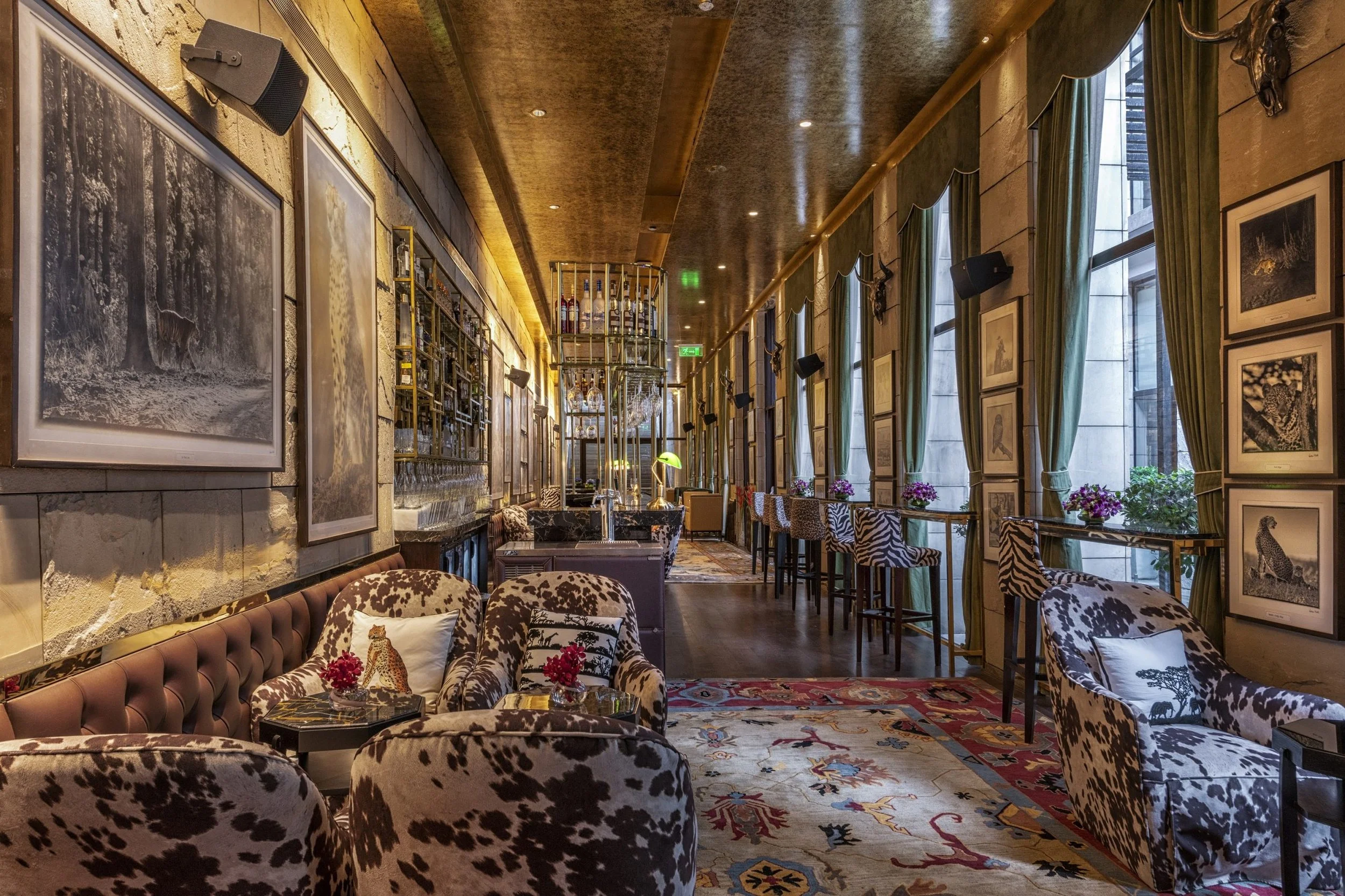 Interior of a stylish lounge or bar with animal-print chairs, framed black-and-white photos on the walls, green curtains, a bar area in the background, and a colorful rug on the floor.