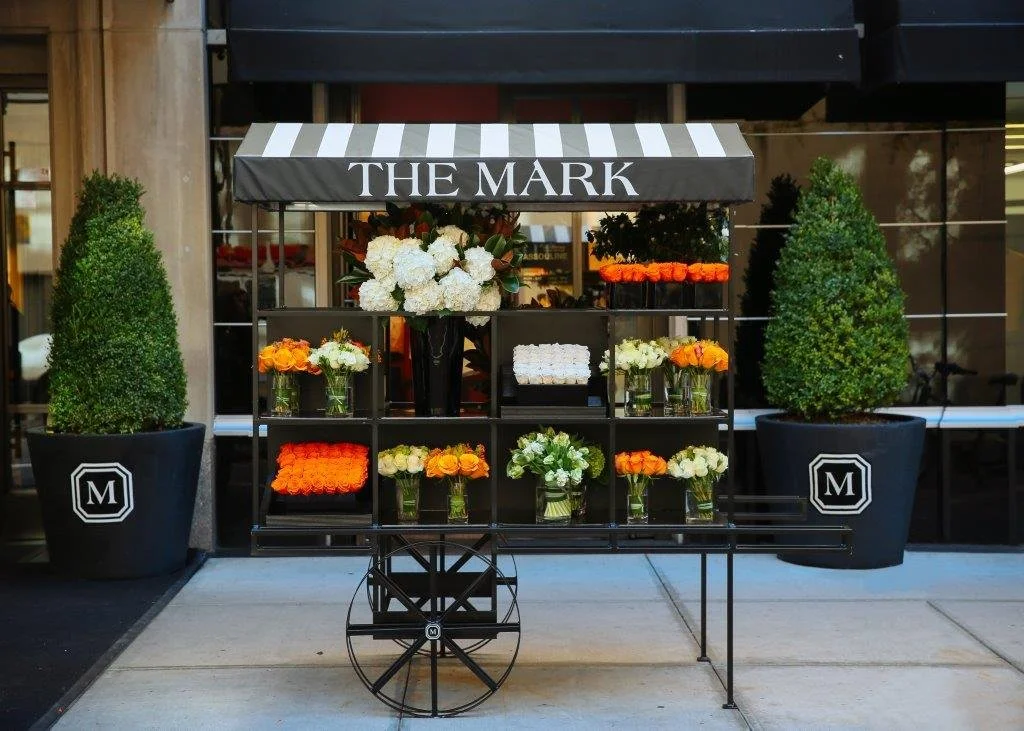 Flower cart with white, yellow, and orange flowers outside a shop called 'The Mark'; two large potted green bushes on either side.