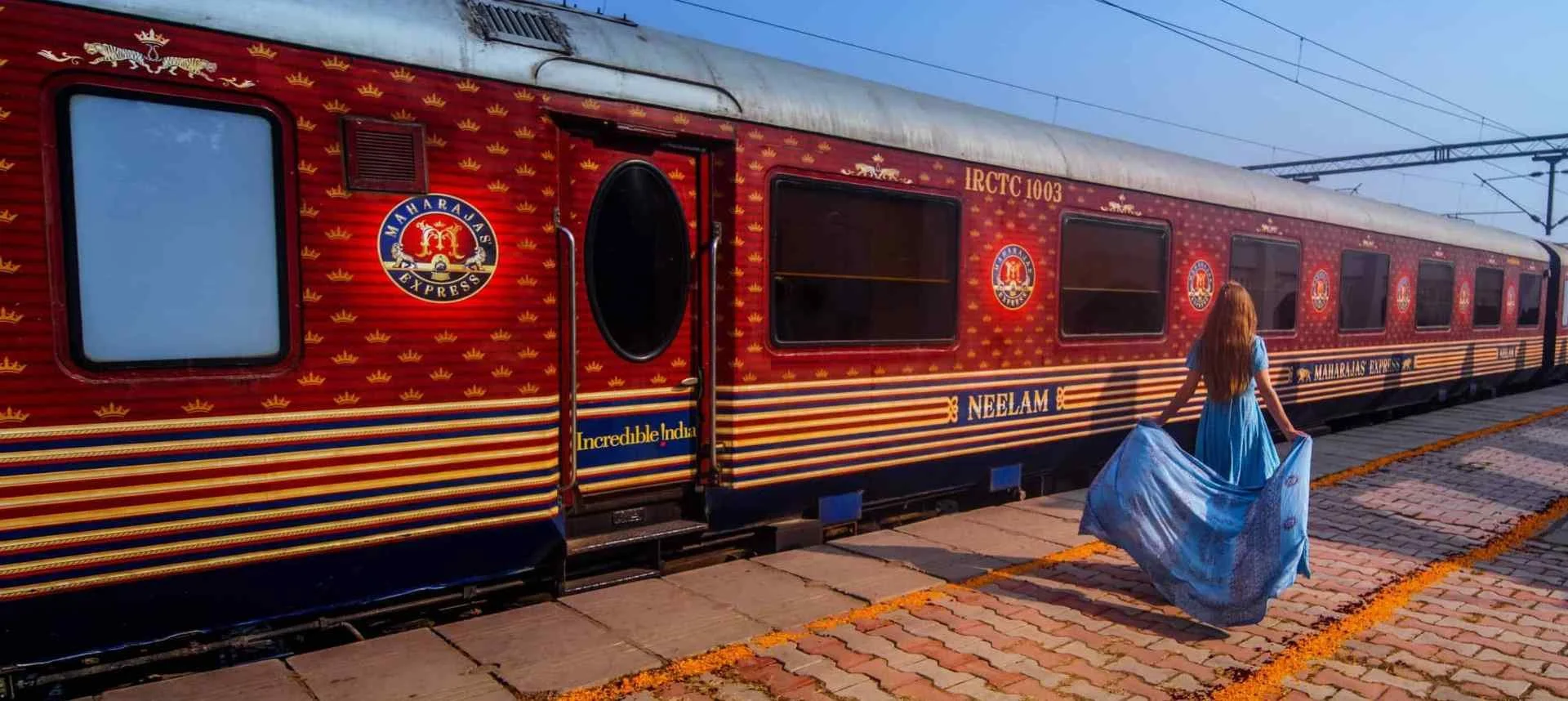 A woman in a blue dress walking along a red train on a station platform. The train has Maharajas Express branding and is decorated in red with gold and blue accents. The station has paved brick flooring and overhead electric wires.