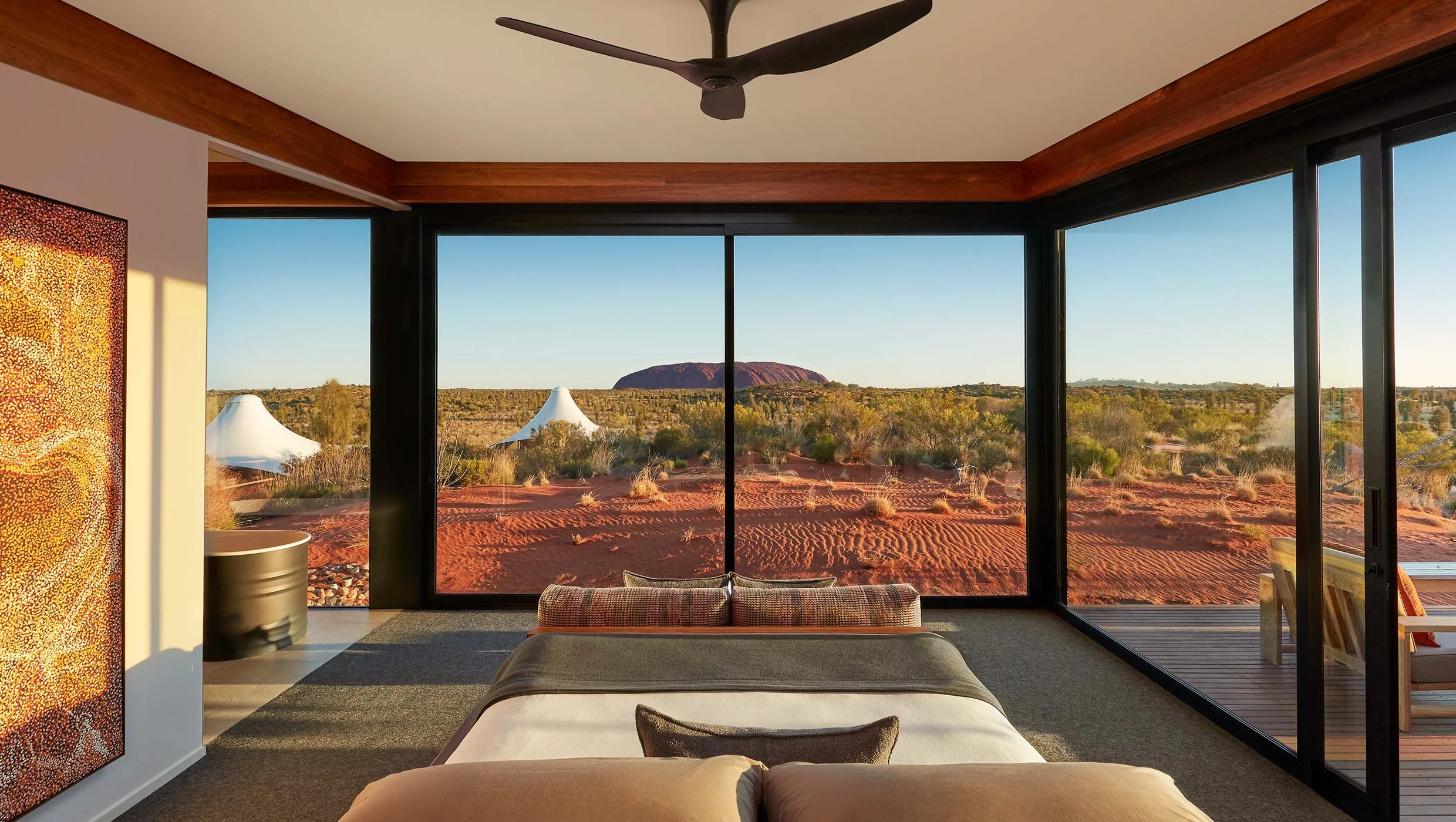 Interior of a modern room with large floor-to-ceiling windows showing a desert landscape with red sand, bushes, and a large rock formation in the background. The room has a bed, a ceiling fan, a side table, and a colorful painting on the wall.