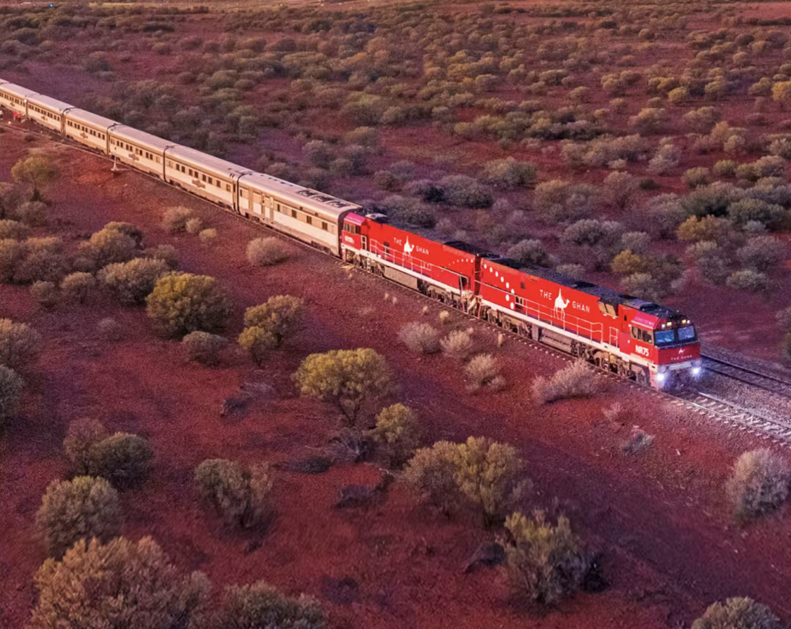 A red train with the words "The Ghan" on the side travels through a desert landscape with bushes and small trees, under a pink and purple sky.
