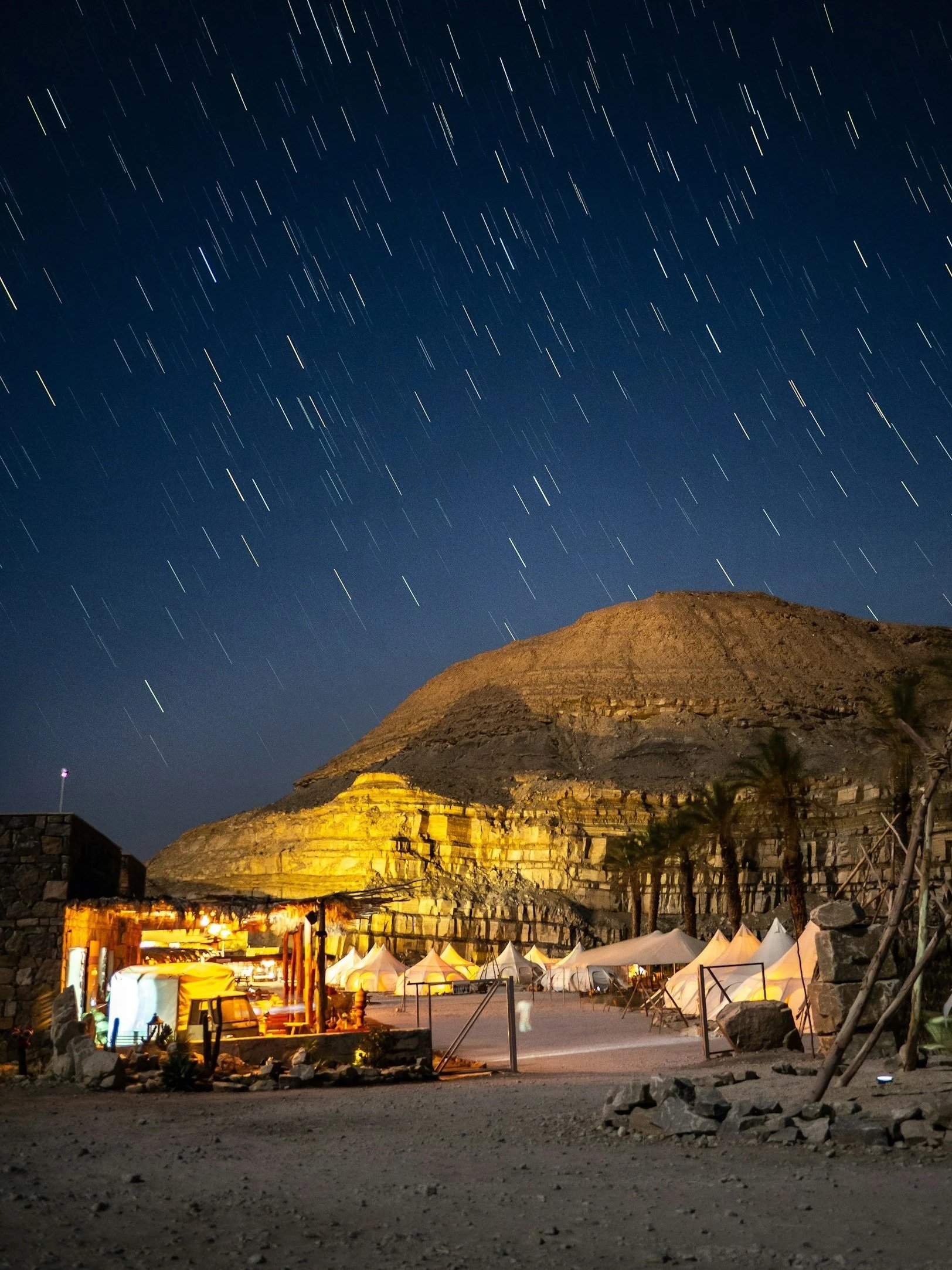 Star trails over a desert landscape with tents and a mountain in the background at night.