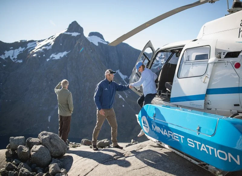 Three men standing on rocky terrain near a helicopter marked with Minaret Station logo, with a mountain and snow patches in the background. One man is shaking hands with another as a third looks on.