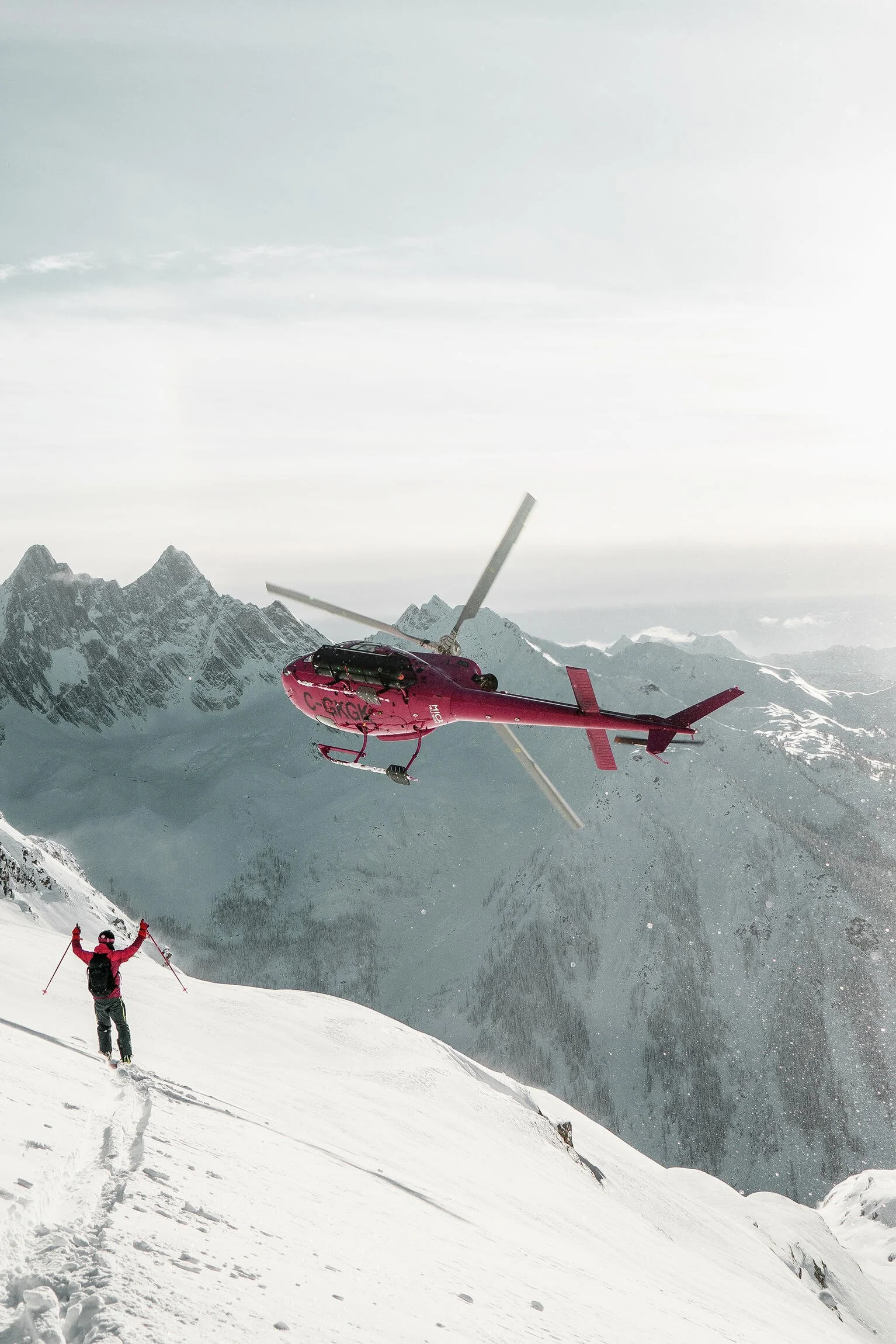 A person in pink gear skiing on snow-covered mountains while a helicopter flies nearby in a mountain landscape.