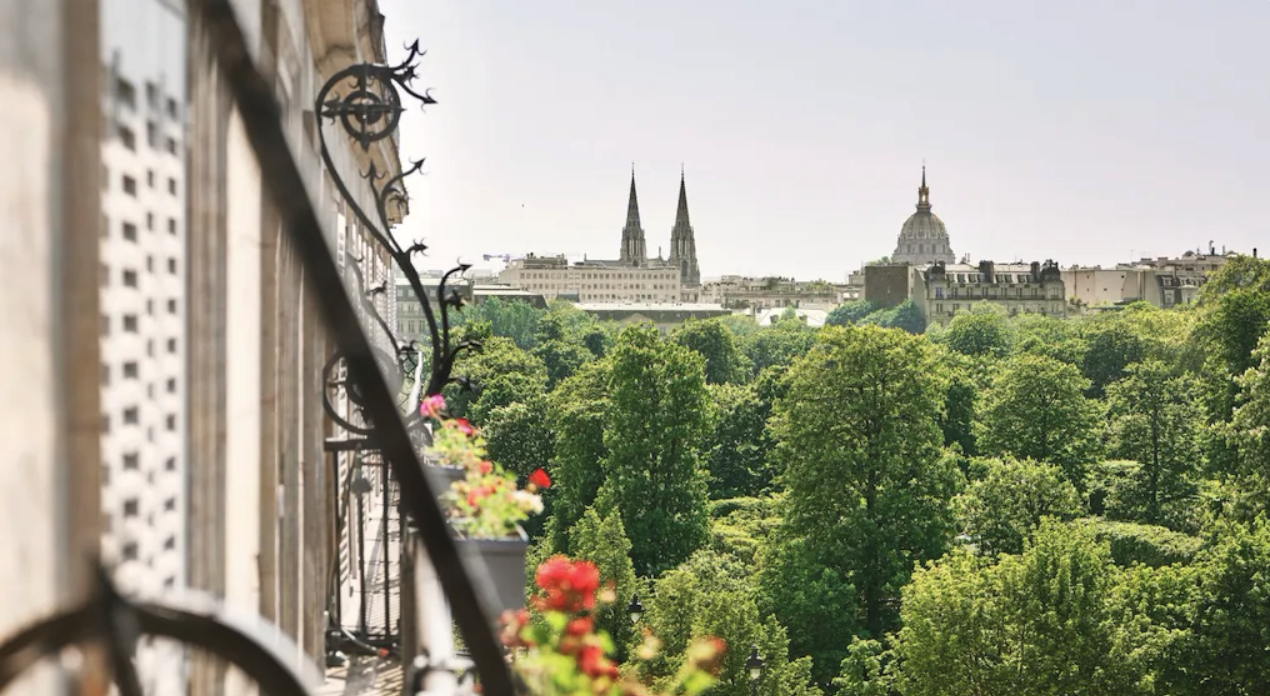 View of a city skyline with church spires and domed buildings over green trees, seen from a balcony with ornate black metal railings and flower boxes with pink and red flowers.