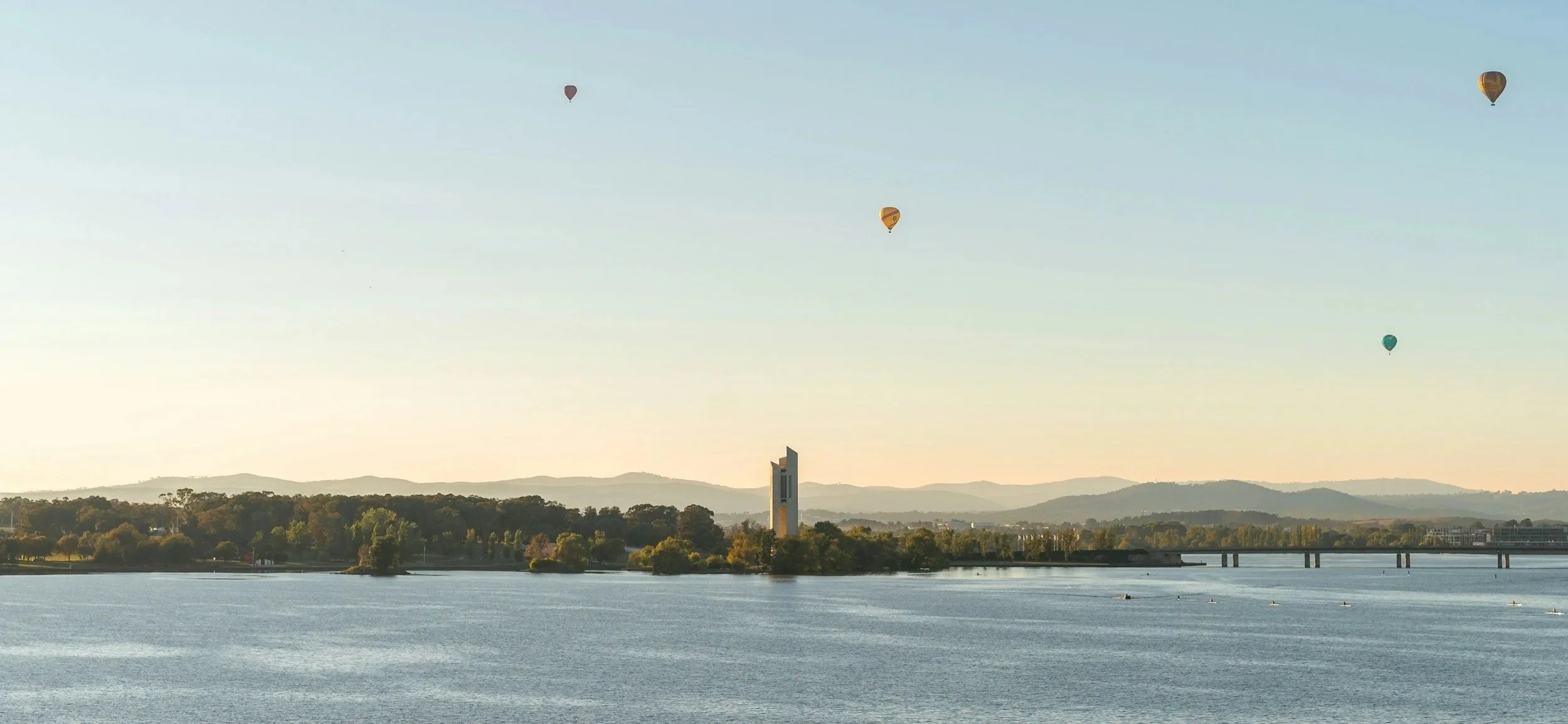 Four hot air balloons floating in the sky over a large body of water with a distant bridge, trees, and hills in the background during daylight.