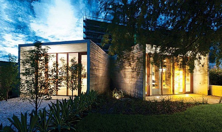 Modern concrete house with large glass windows illuminated from inside, set in a landscaped yard during dusk.