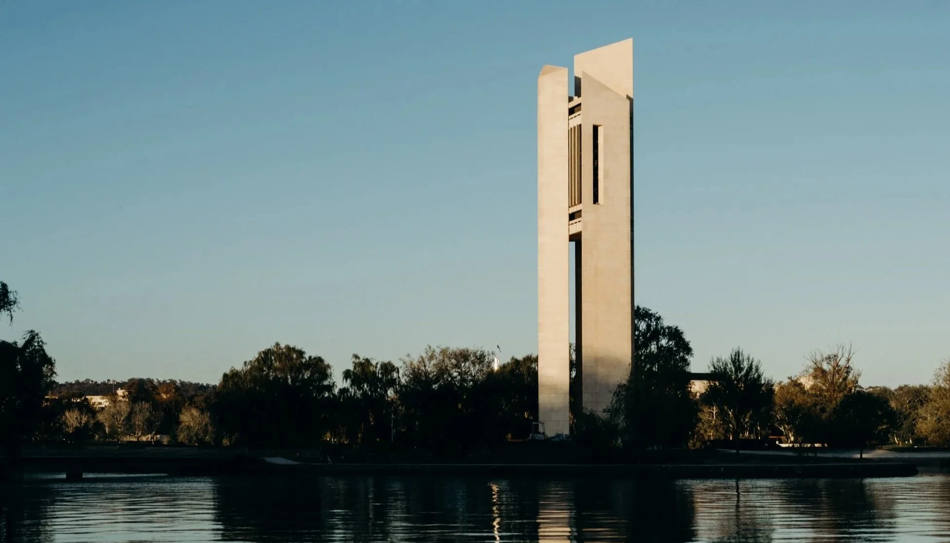 A tall, modern monument or building with geometric shapes stands above trees near a body of water.