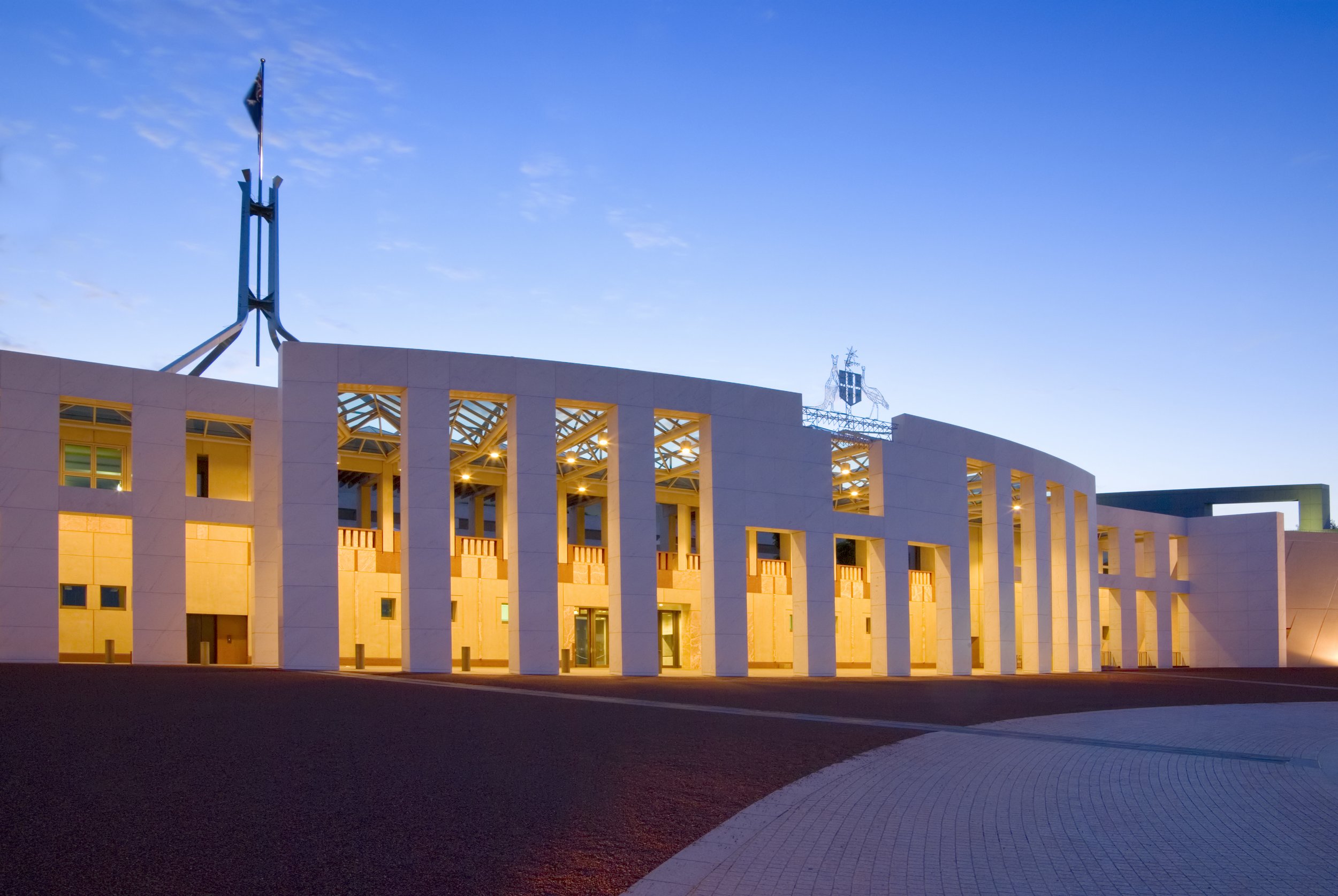 The Australian Parliament House illuminated at dusk with a clear blue sky, modern architecture featuring a white facade, tall vertical windows, and a flag flying at the top of a prominent spire.