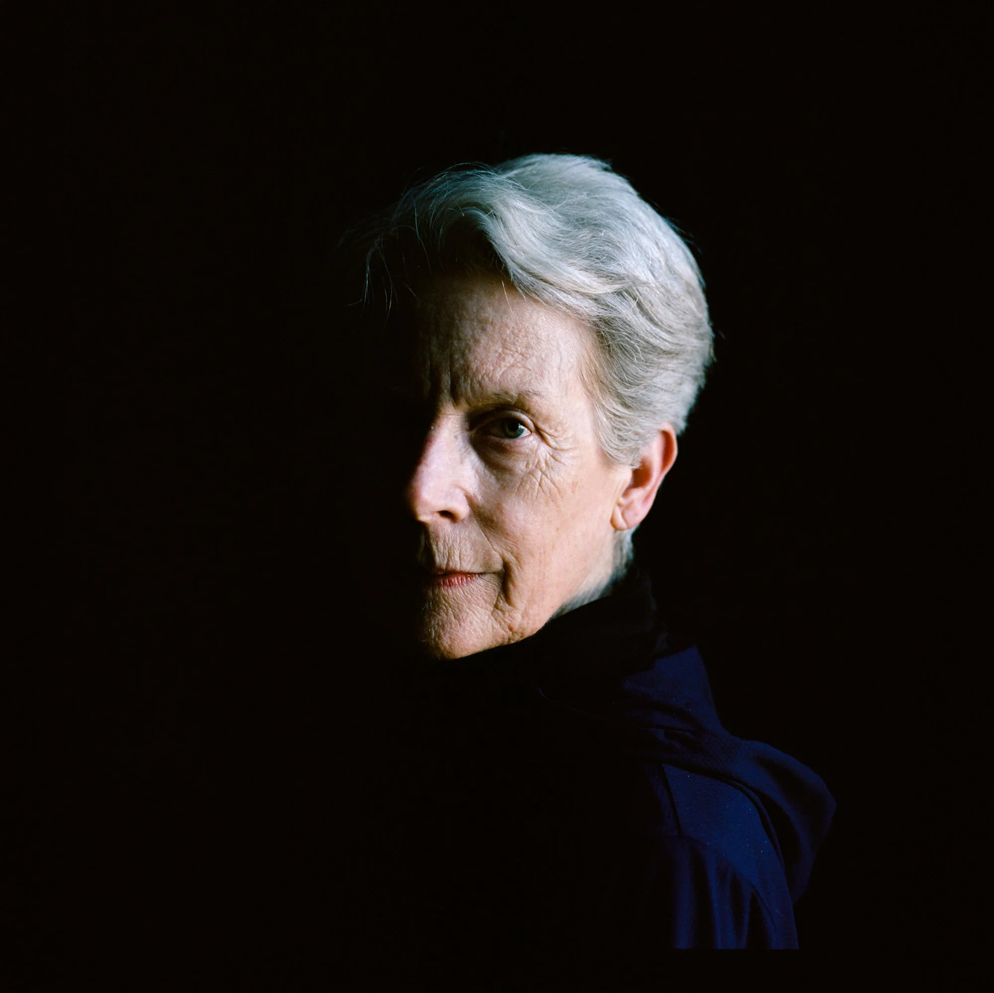 Half-portrait of an elderly woman with gray hair, illuminated from the front left, against a black background, wearing a dark top.