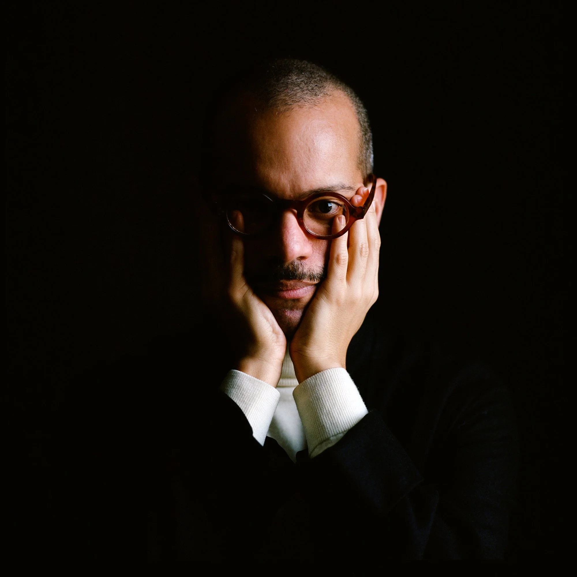 Portrait of a man with glasses, resting his face in his hands, against a dark background.