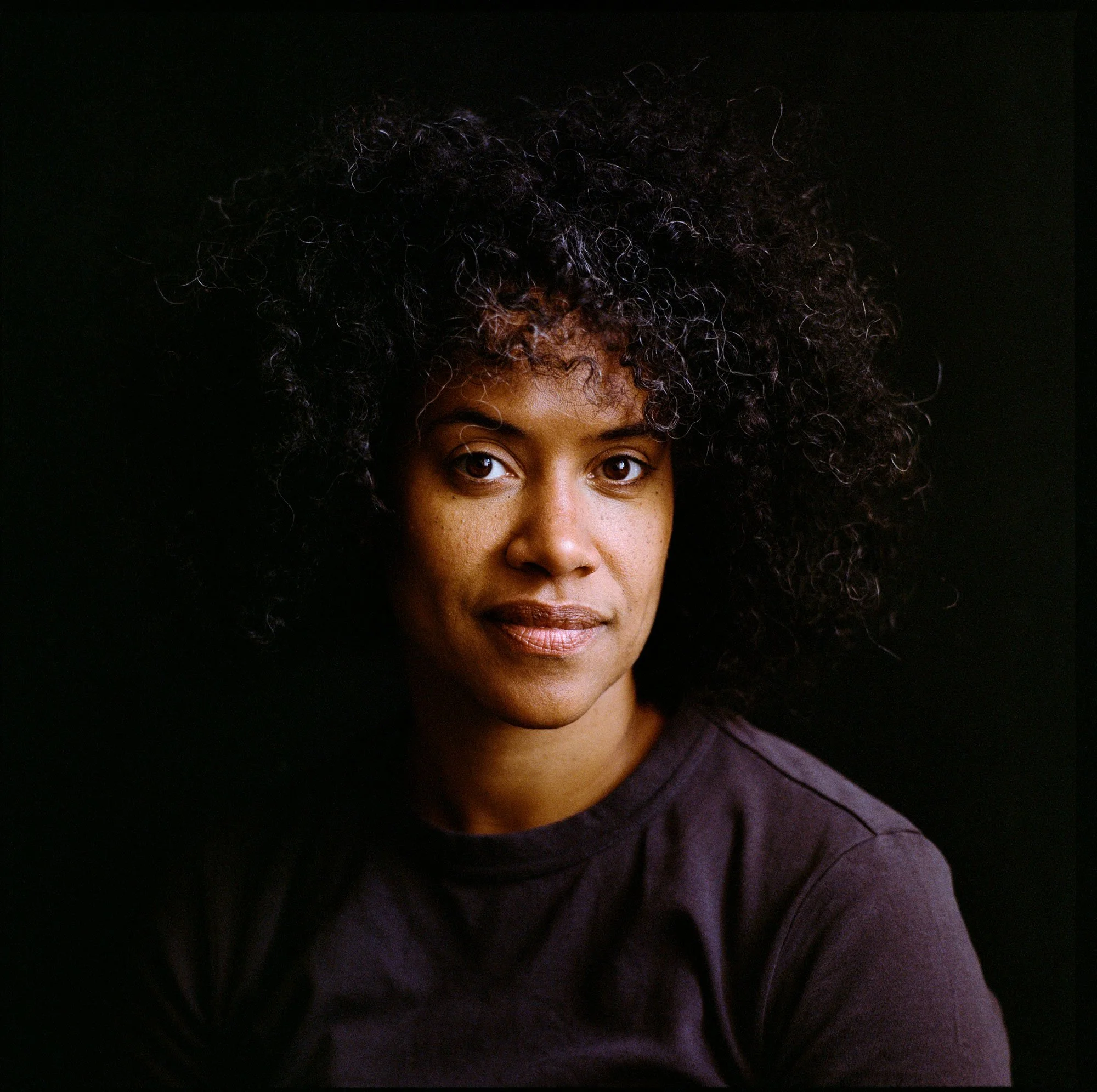 Portrait of a woman with dark curly hair, wearing a dark t-shirt, against a black background.