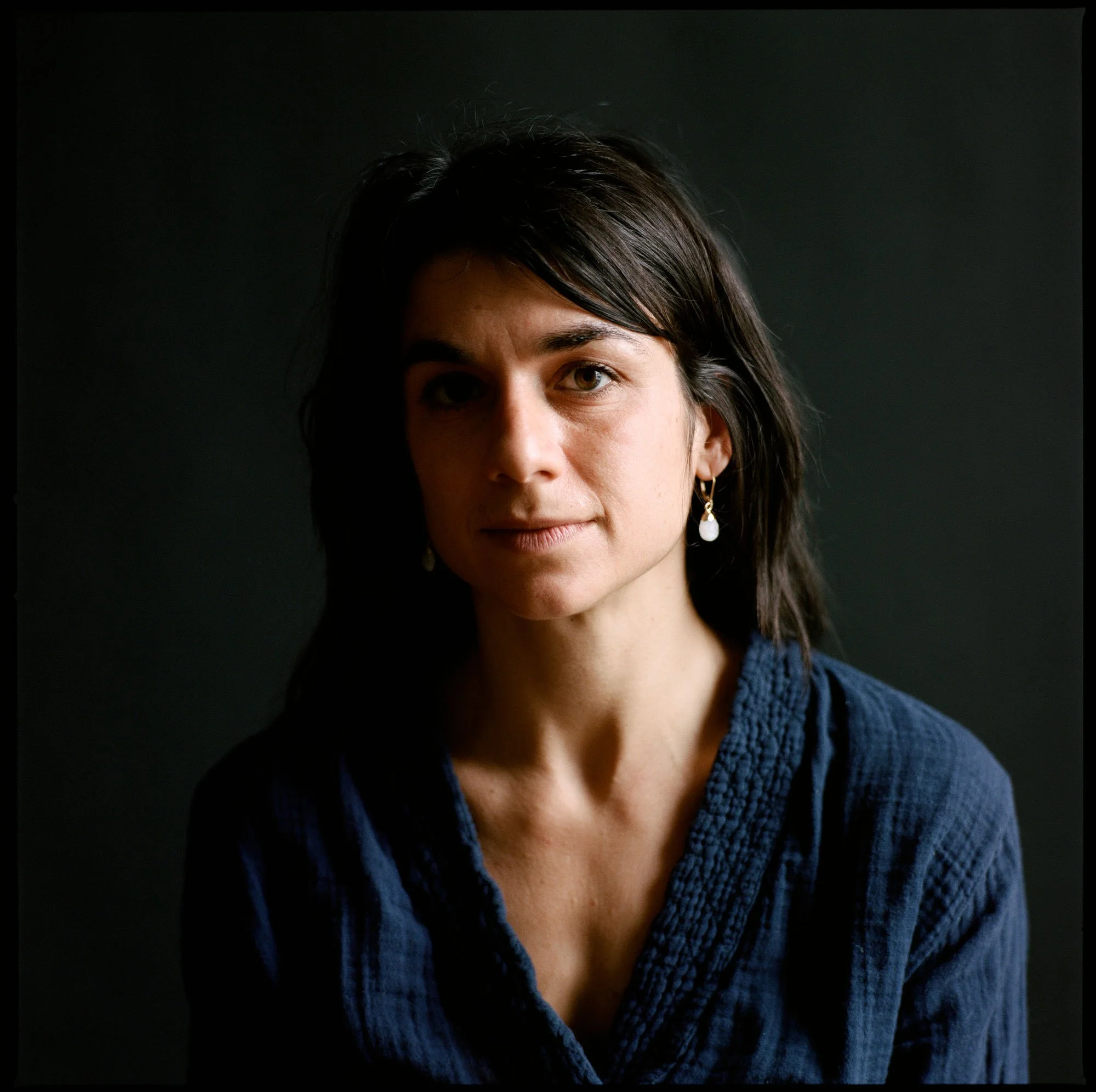 Portrait of a woman with dark brown hair, wearing earrings and a blue textured blouse, against a dark background.