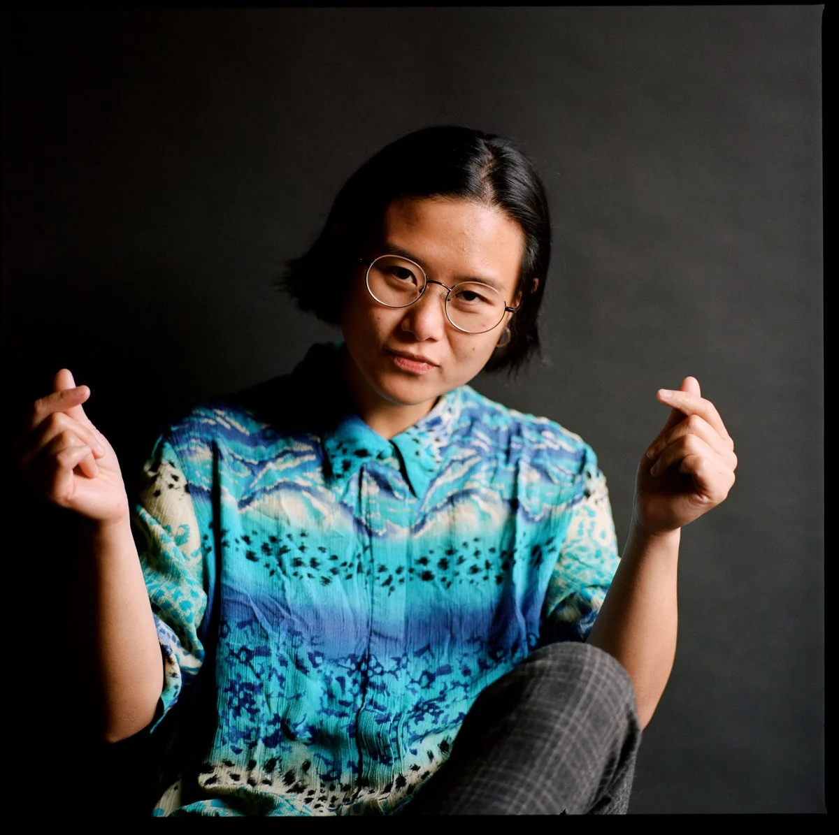 A person with short black hair, glasses, and light skin, wearing a colorful blue, green, and black patterned shirt, making a finger heart gesture with both hands, sitting against a dark background.