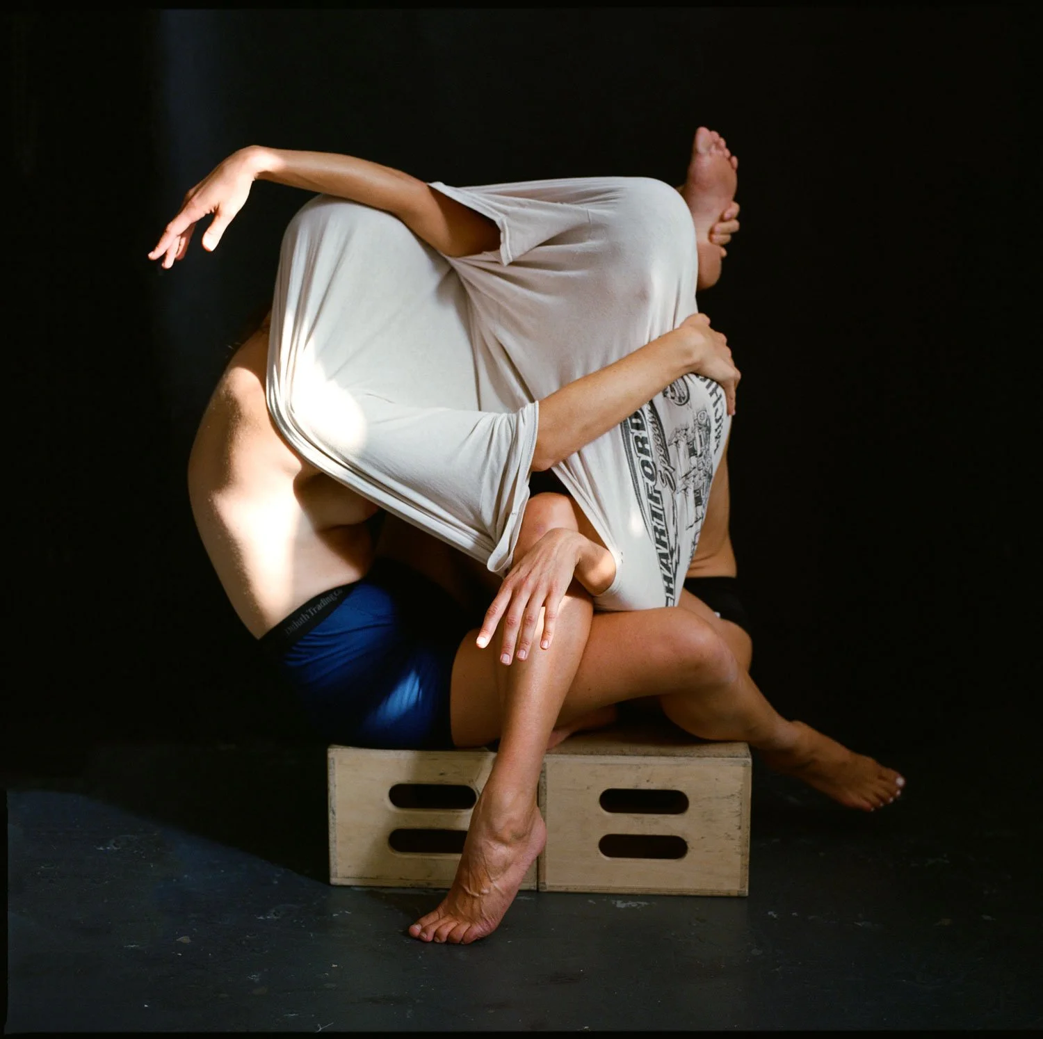 Two women engaged in a modern dance or performance art, intertwined and balancing on a wooden box, with a dark background.
