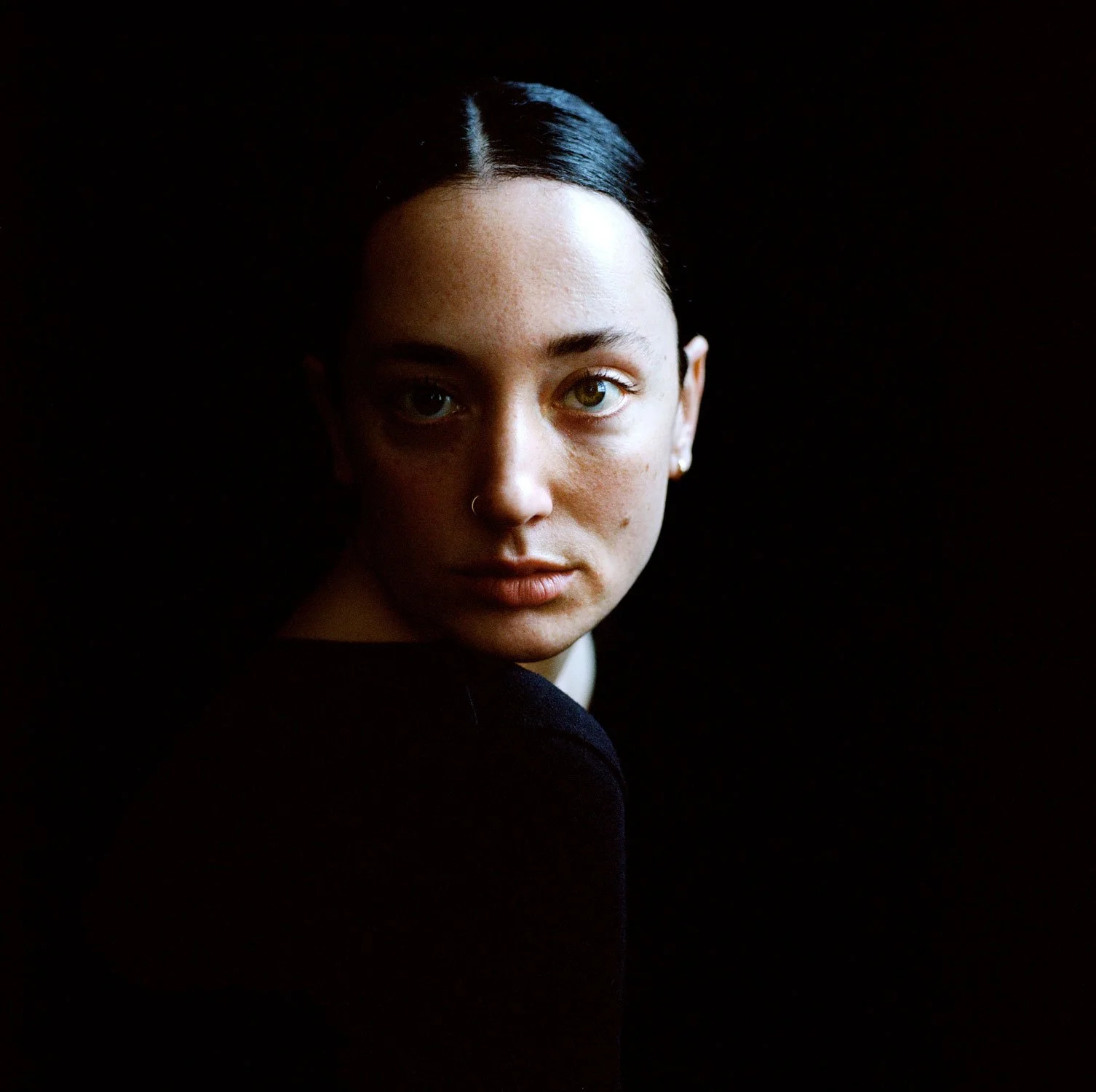Portrait of a young woman with dark hair slicked back, looking directly at the camera against a black background.