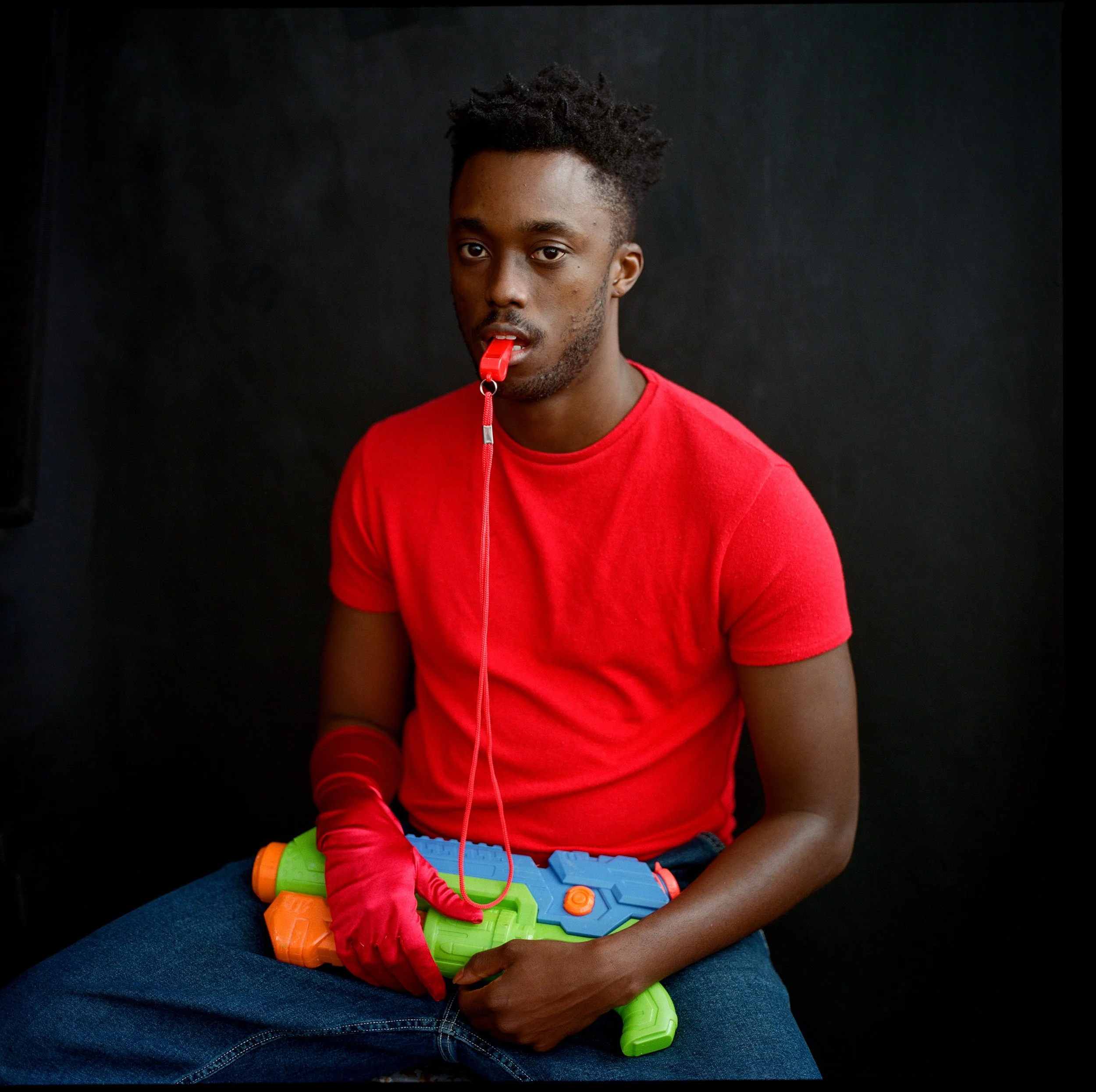 A young man in a red t-shirt with a toy water gun and a whistle in his mouth, sitting against a dark background.