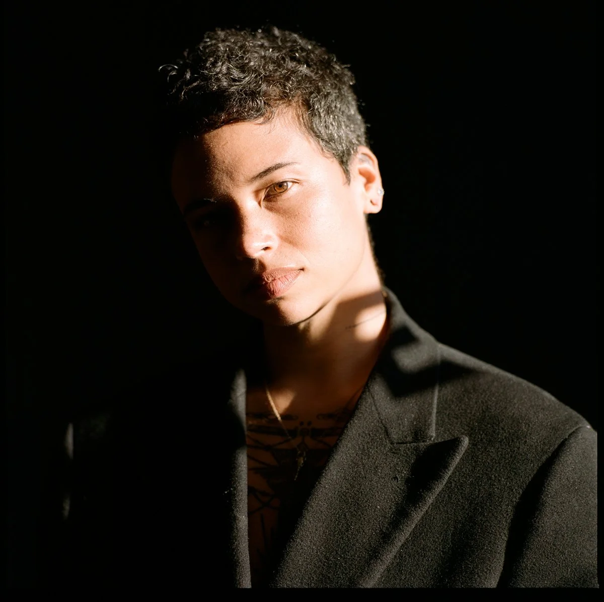 Portrait of a young woman with short curly hair, wearing a black blazer and a necklace, illuminated by dramatic lighting against a black background.