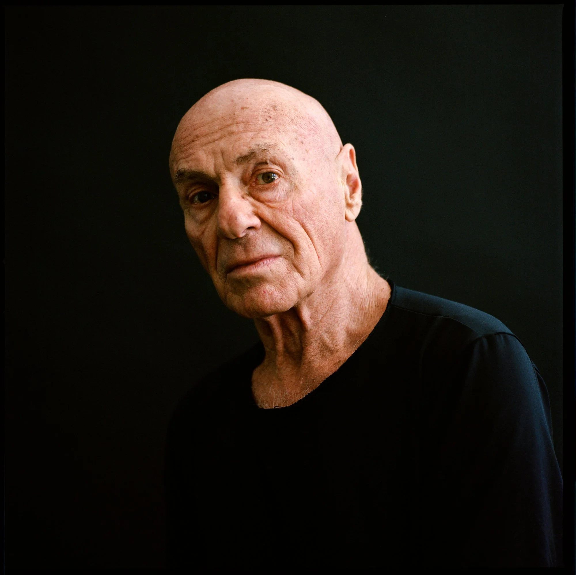 Portrait of an elderly bald man wearing a black shirt against a dark background.