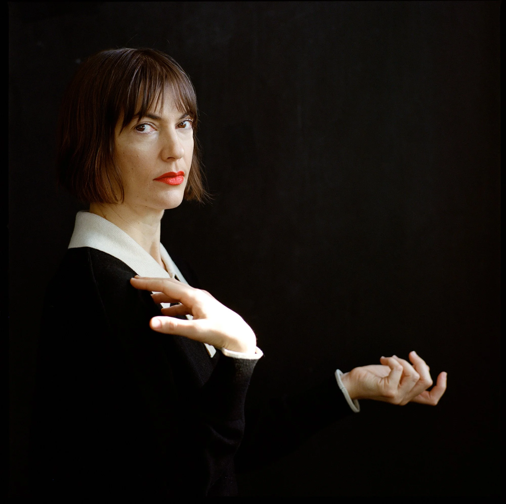A woman with short brown hair, red lipstick, wearing a black and white outfit, standing against a black background, looking at the camera with her hands positioned near her shoulders.