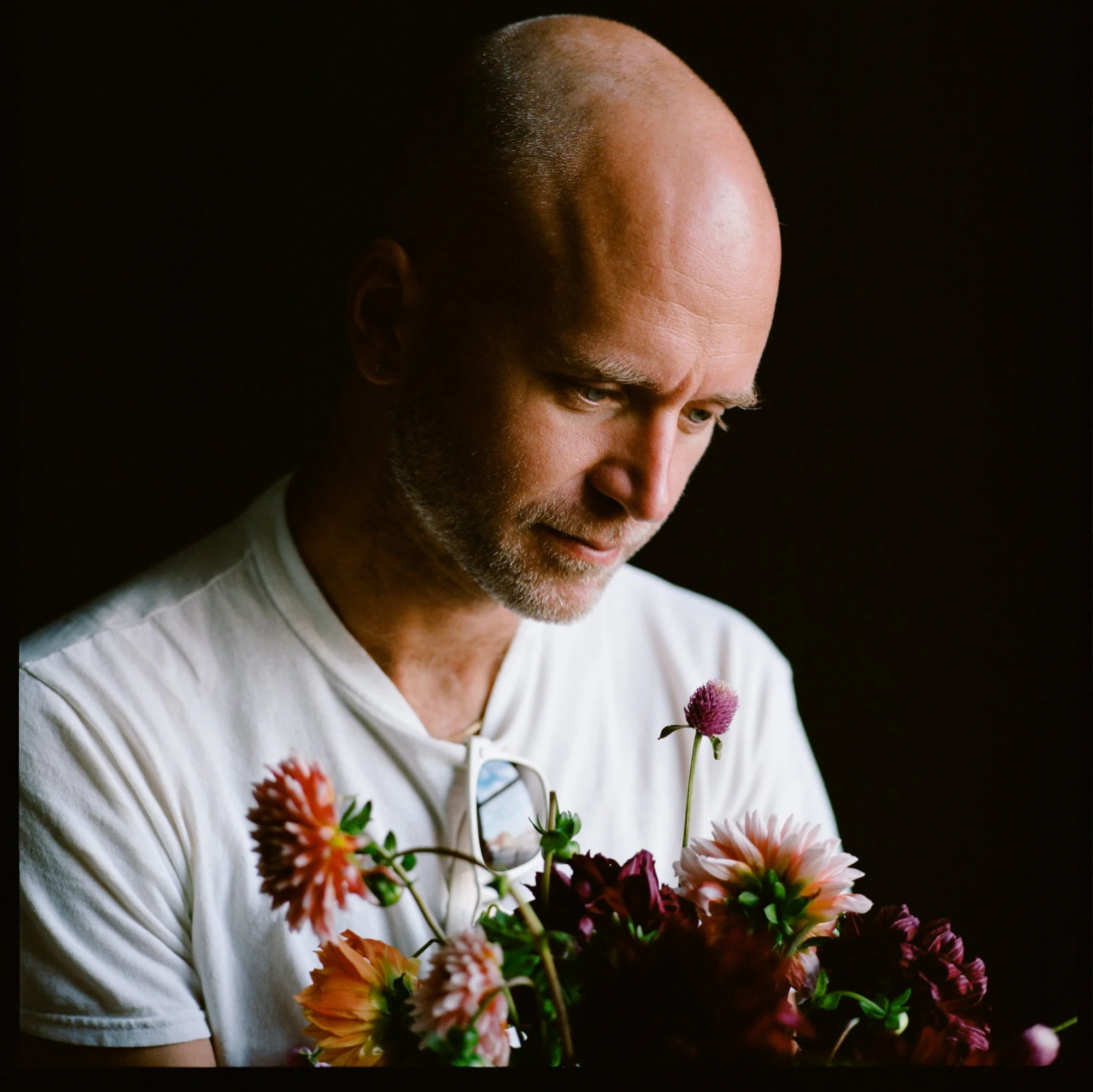 A bald man with a beard looking down at a bouquet of various colorful flowers against a black background.