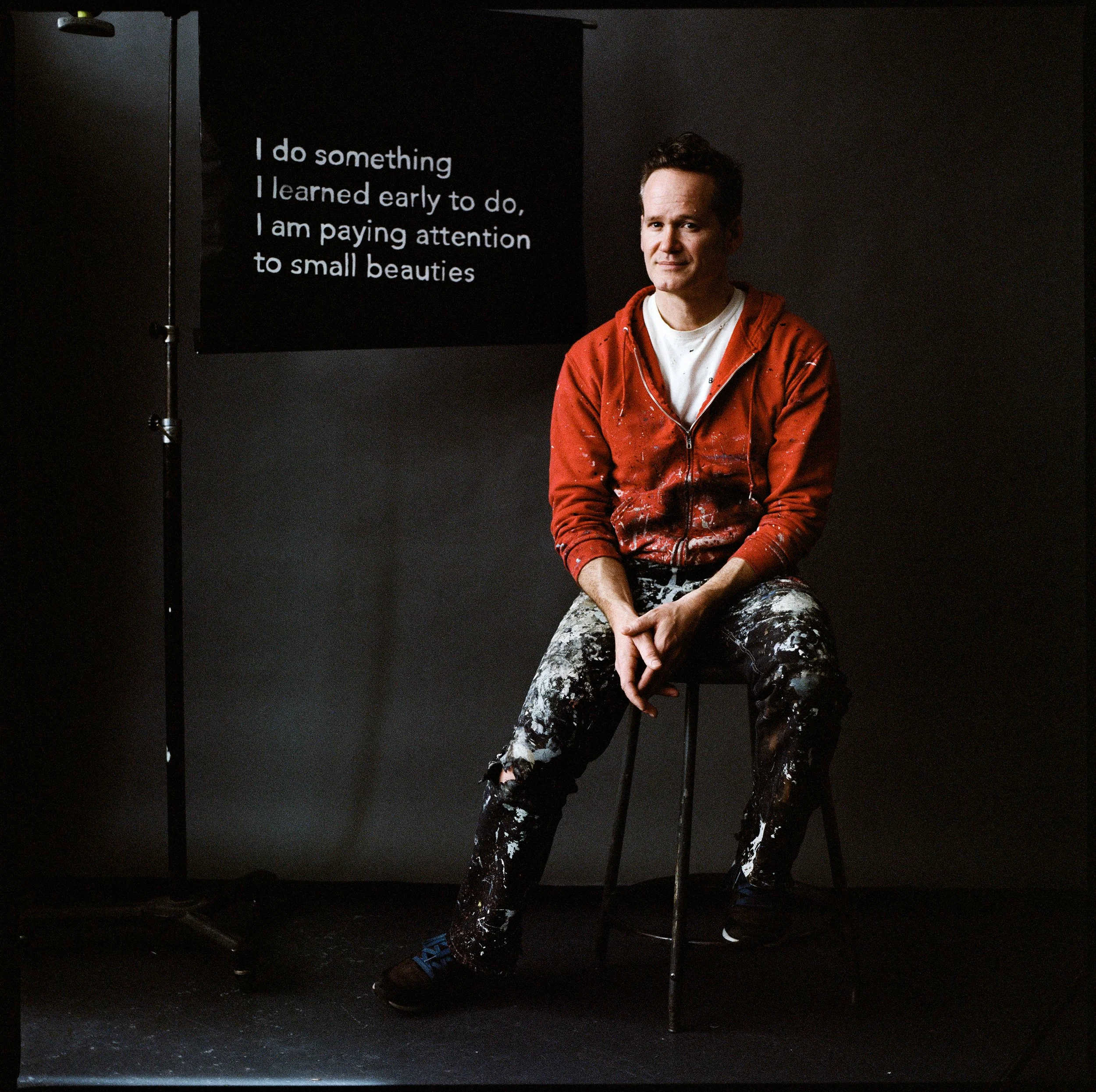 A man in a red hoodie and paint-splattered black pants sits on a stool in front of a black background. A sign with white text reads: 'I do something I learned early to do, I am paying attention to small beauties.'