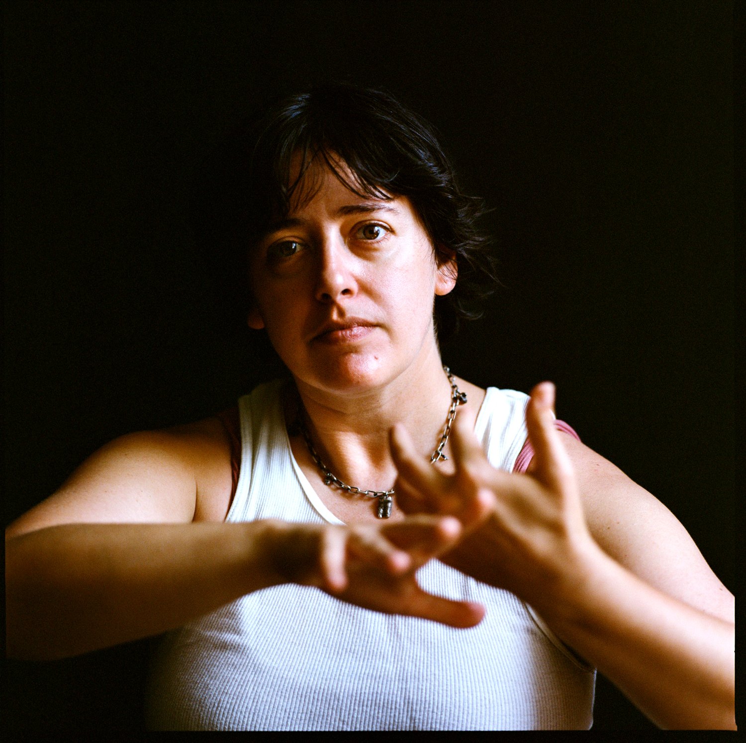 A woman with short dark hair wearing a white tank top and a chain necklace, reaching out with both hands against a dark background.