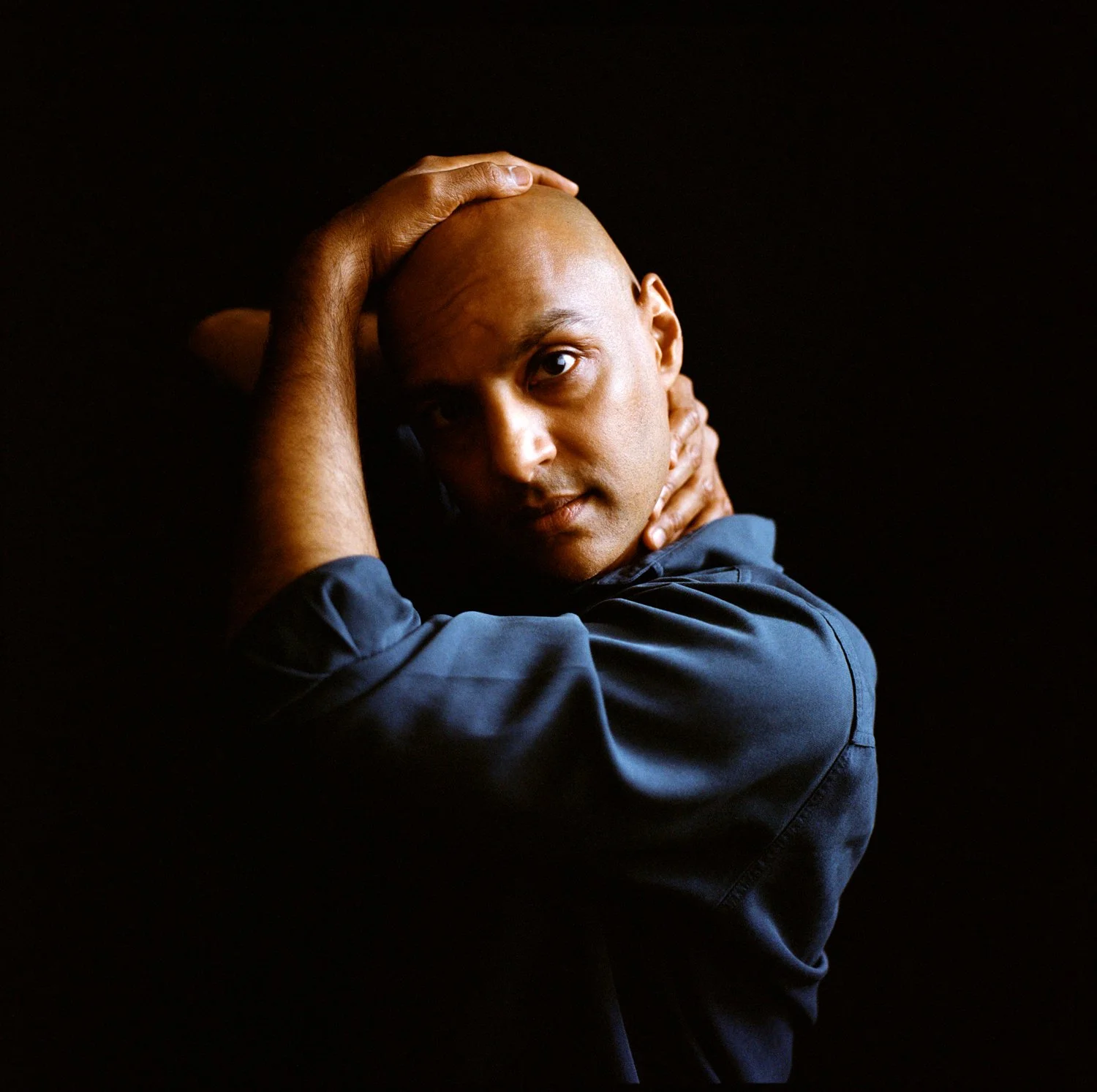 A man with a shaved head posing against a dark background, touching his head and neck with both hands, wearing a dark blue shirt.