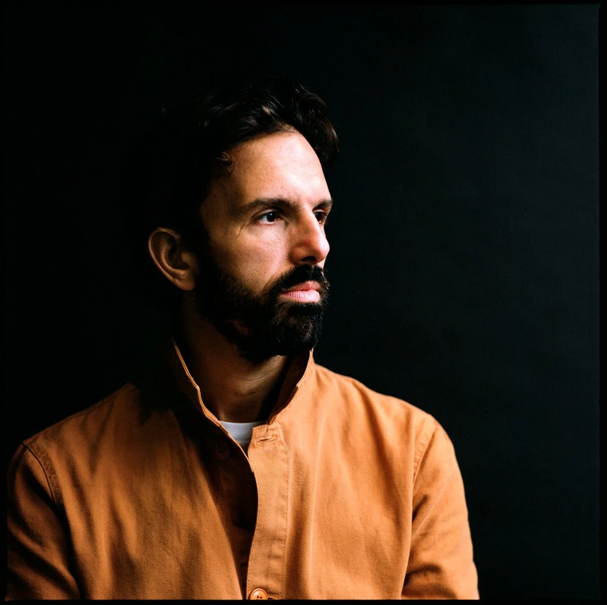 A man with dark hair, a beard, and light skin looking to the right, wearing a brown shirt against a black background.