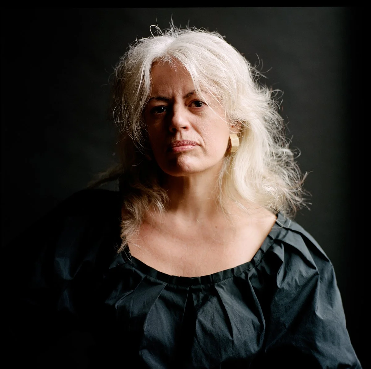 A portrait of an older woman with long, wavy, white hair, wearing a black top, against a dark background.