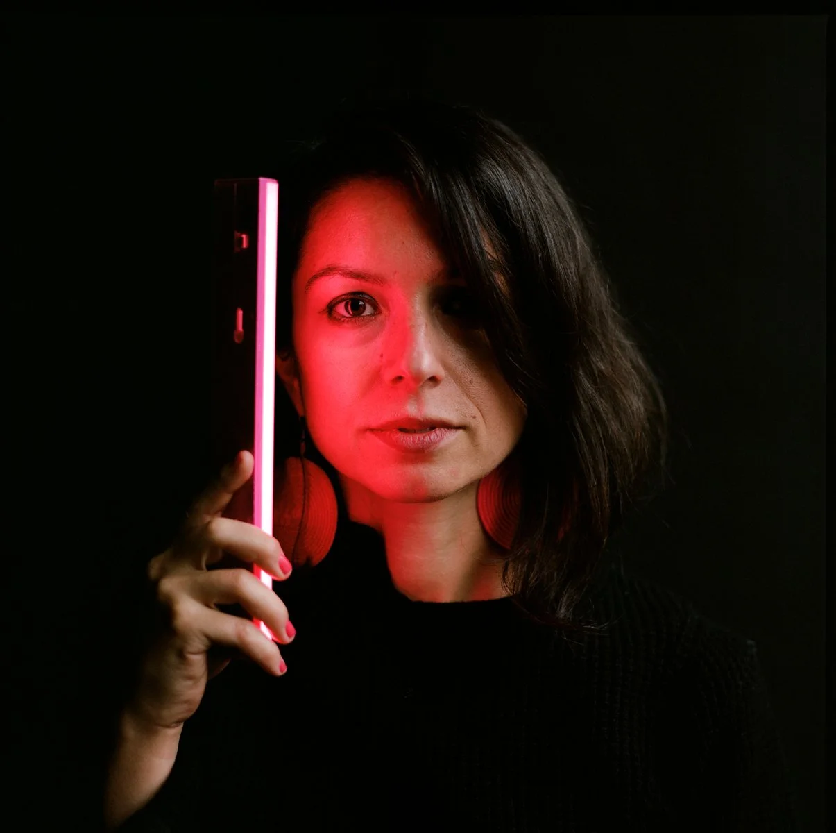 A woman with dark hair wearing red headphones holds a neon pink and white light stick next to her face against a black background.