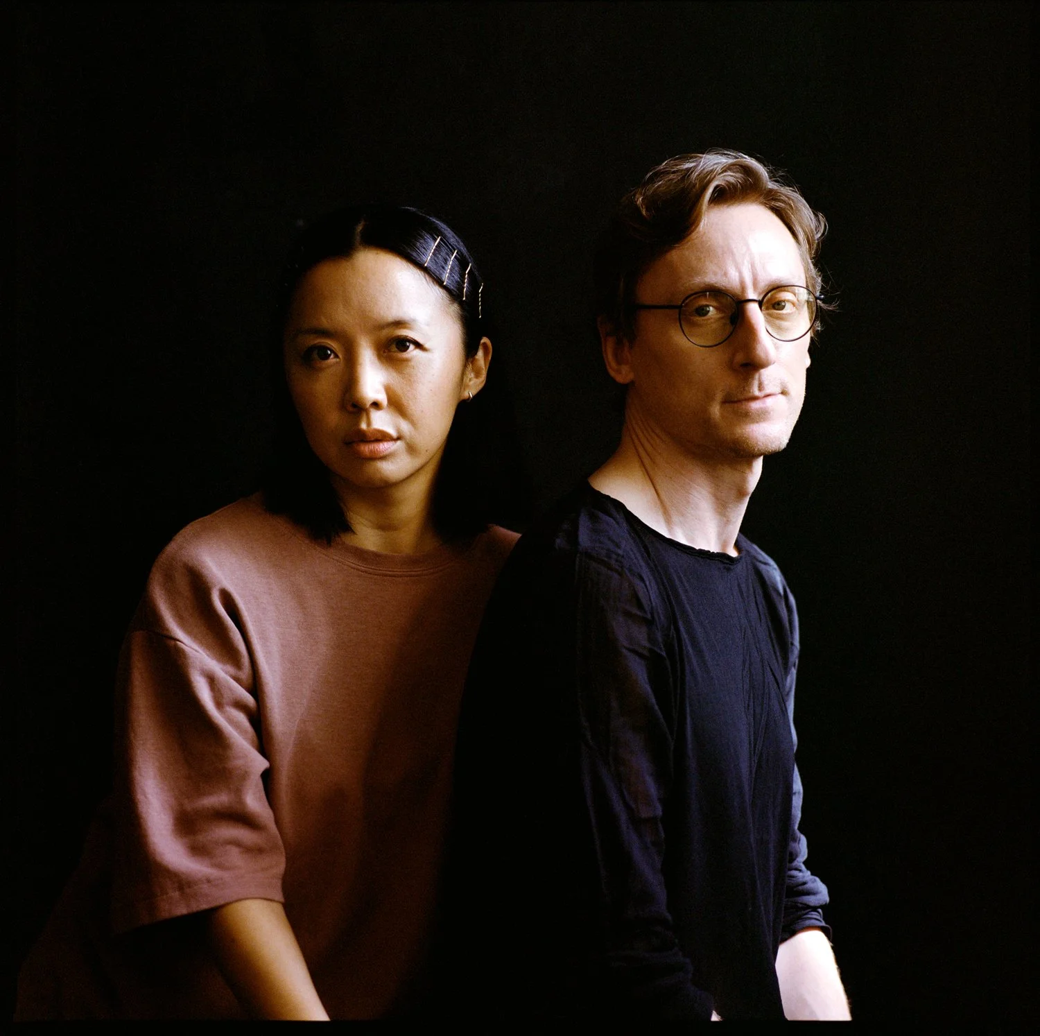 A woman with black hair (left) wearing a pink shirt and a man with glasses and brown hair (right) wearing a black shirt, both against a black background.