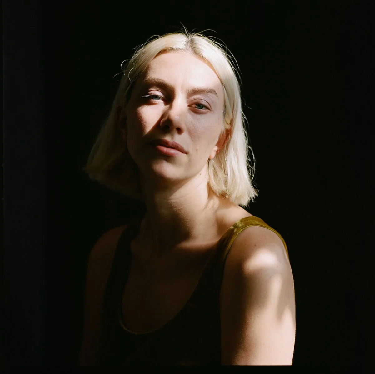 A young woman with platinum blonde hair, wearing a tank top, partially lit by natural light against a dark background.