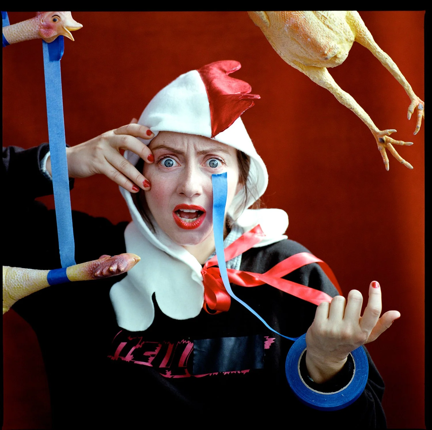 A person dressed in a humorous costume with a chicken hat and a red and white hat, making a surprised expression, with chicken props and strips of blue and pink tape attached to them.