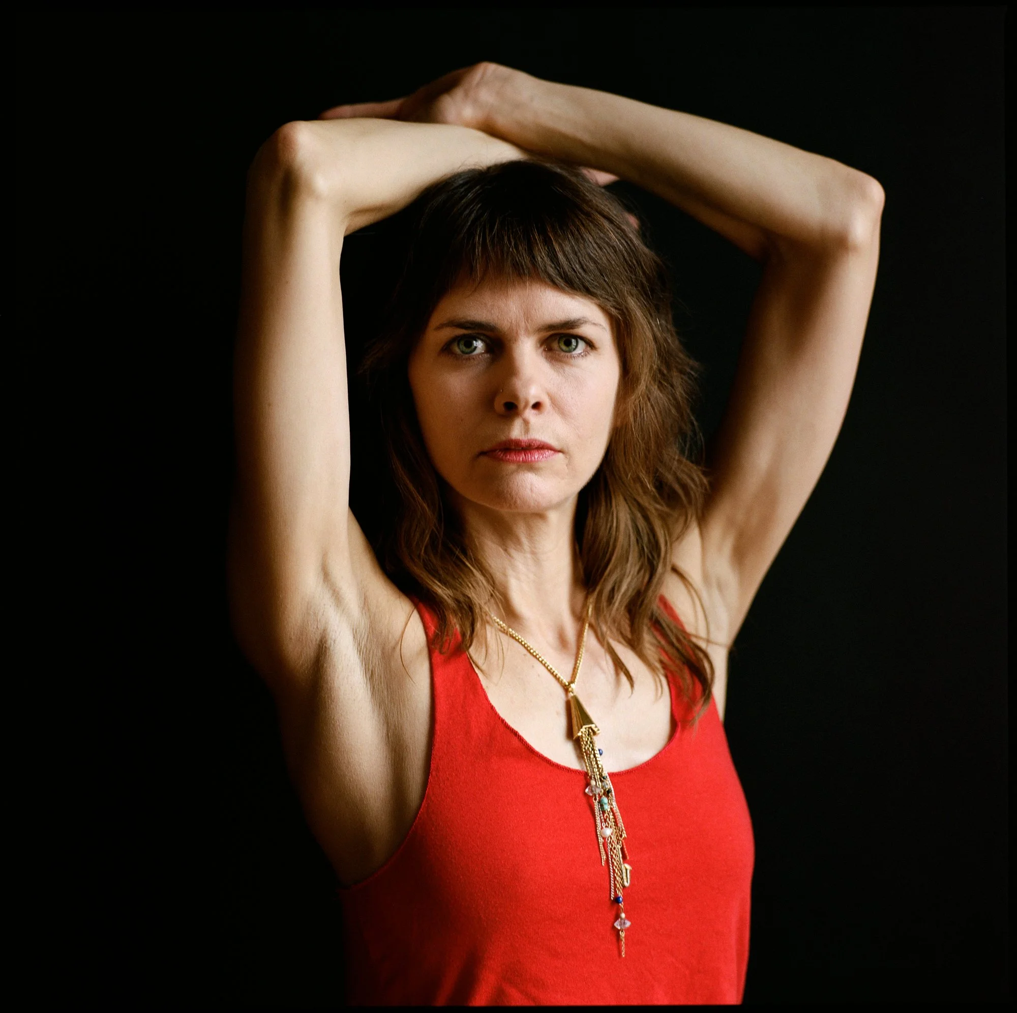 A woman with brown hair and blue eyes wearing a red sleeveless top and a long gold necklace stands against a black background. She has her arms raised above her head, with her hands touching, and looks directly at the camera.