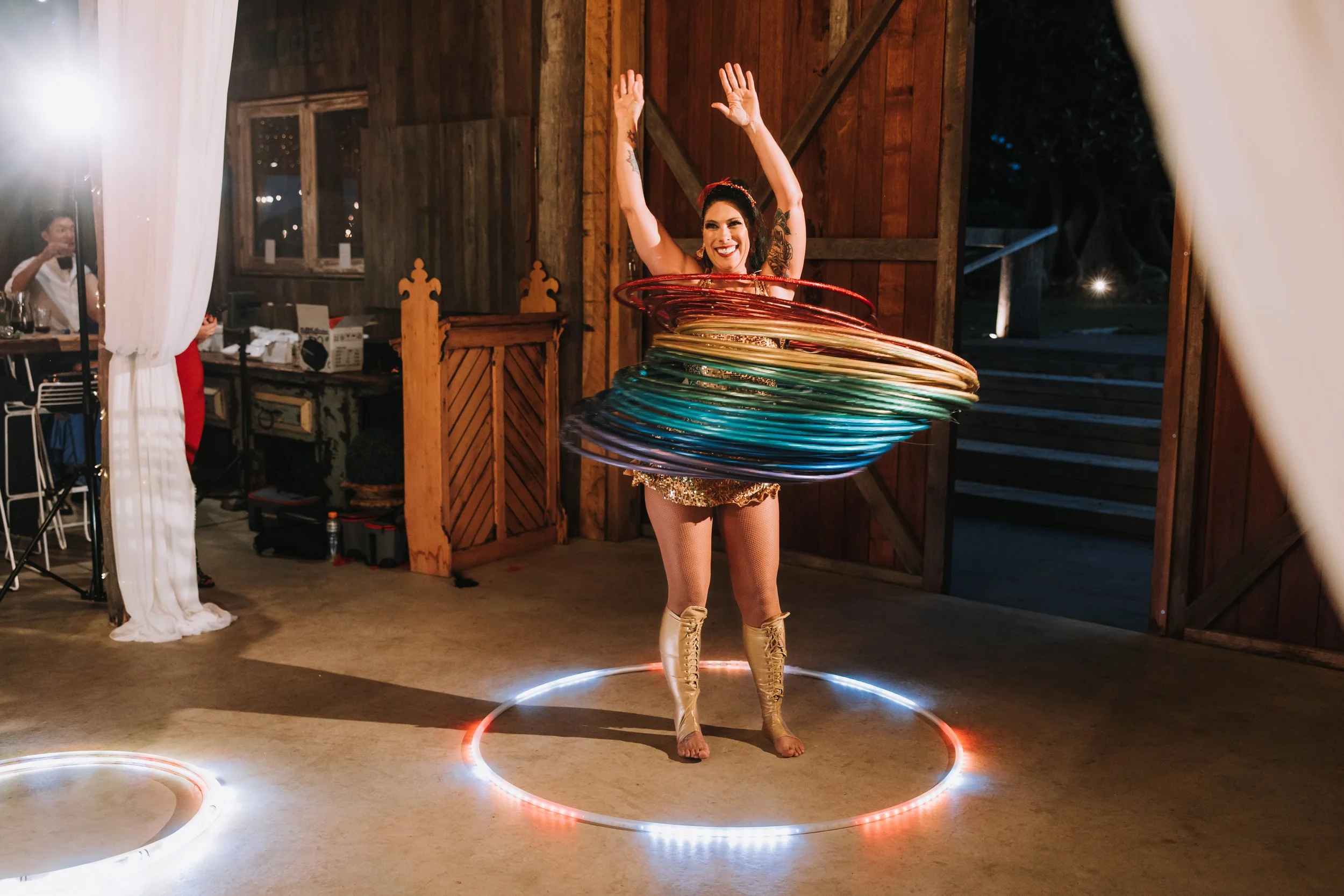 Woman dressed in gold costume spinning colorful hula hoops at a party inside a wooden building.